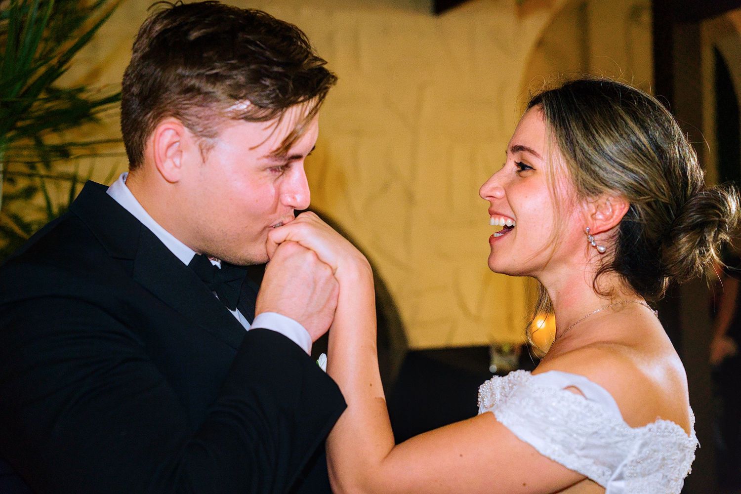 Groom kissing bride's hand while she laughs joyfully during romantic wedding reception dance.