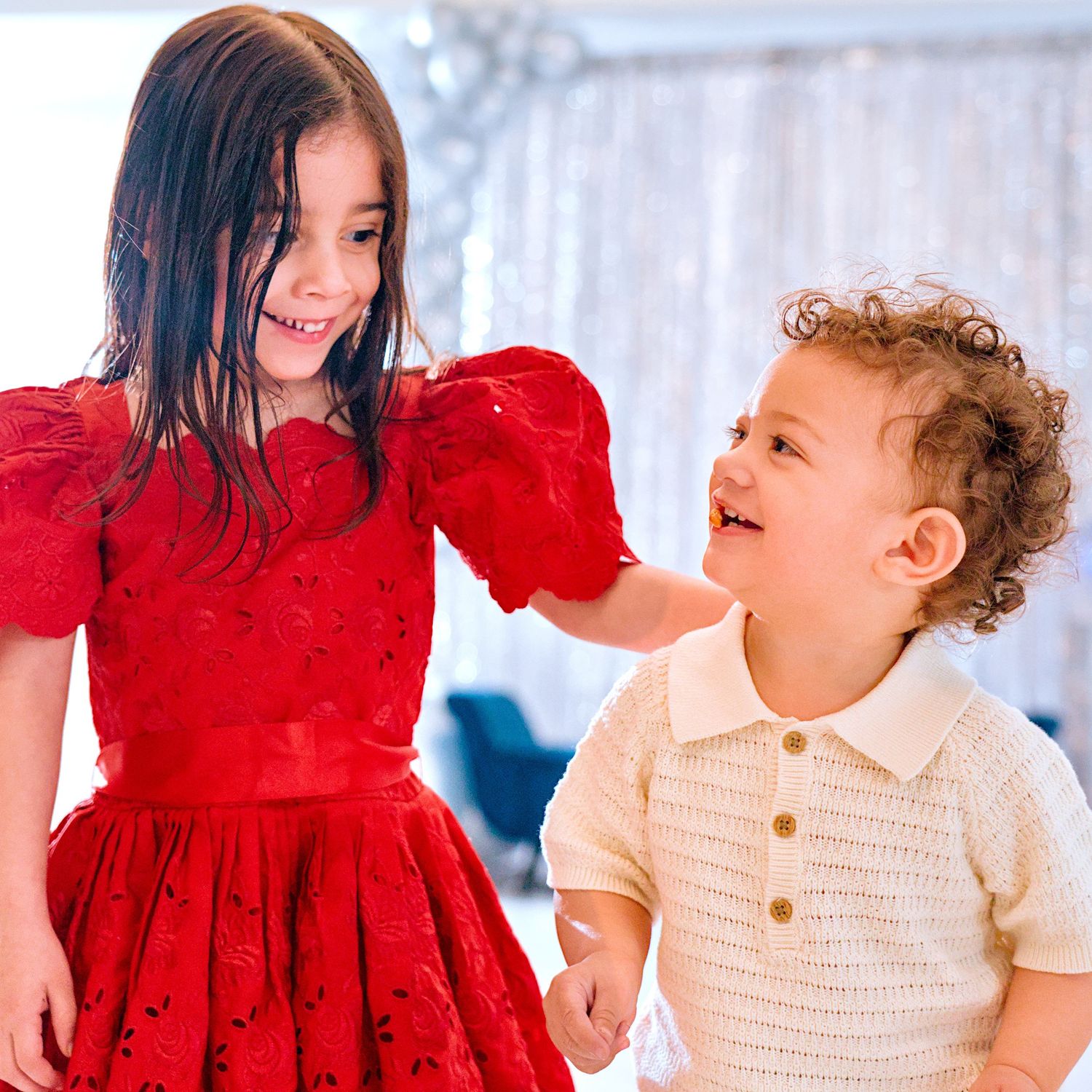 Two young children in festive attire, girl in red dress and boy in cream outfit, laughing together at party.