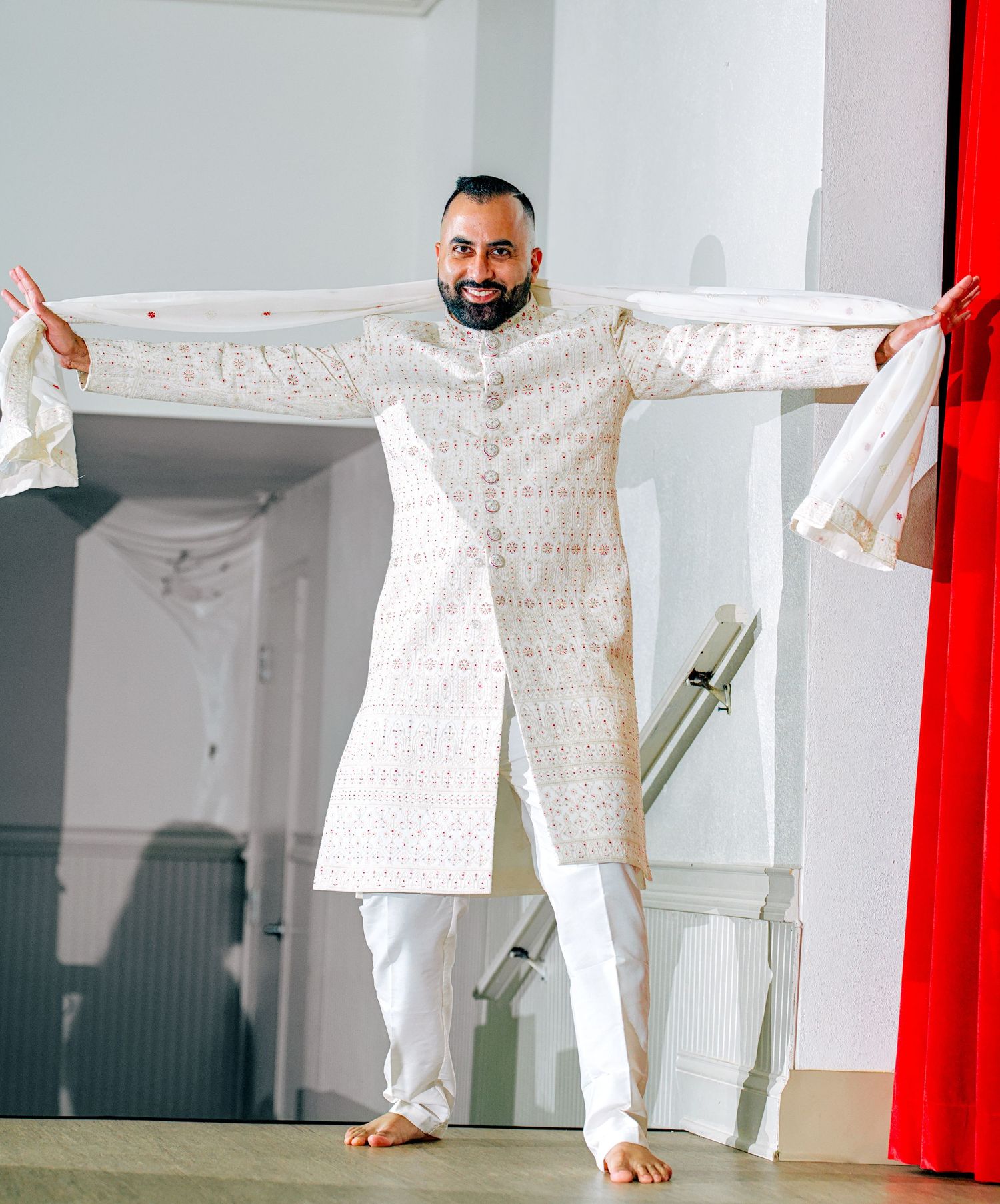 Groom in white embroidered sherwani posing with arms outstretched at Indian wedding.