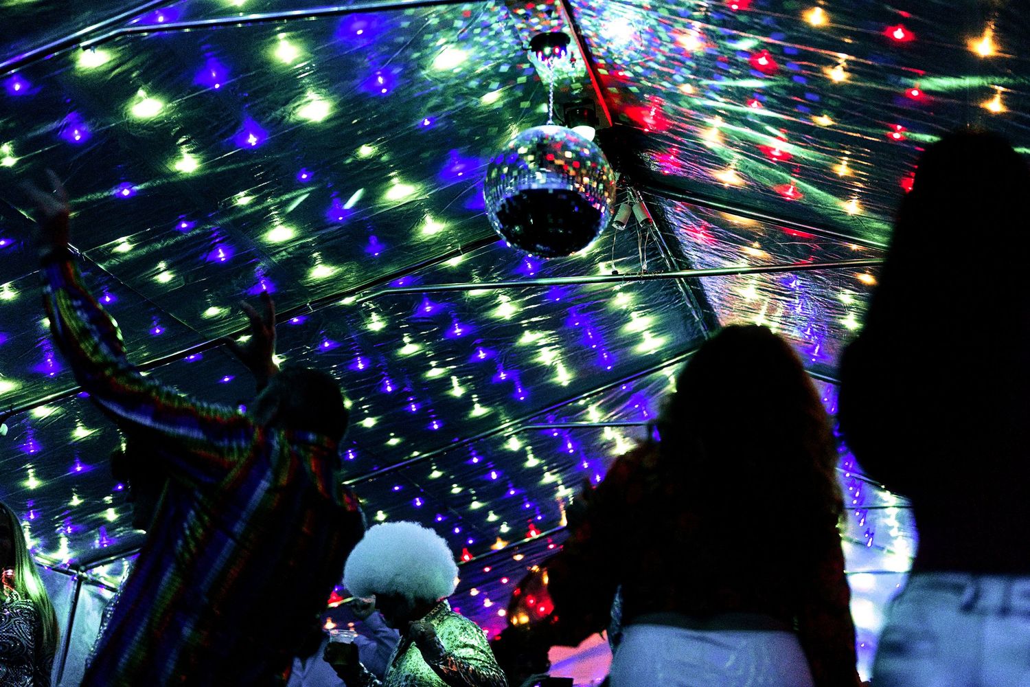 Silhouettes of party guests dancing under a canopy of colorful string lights and a disco mirror ball at night.