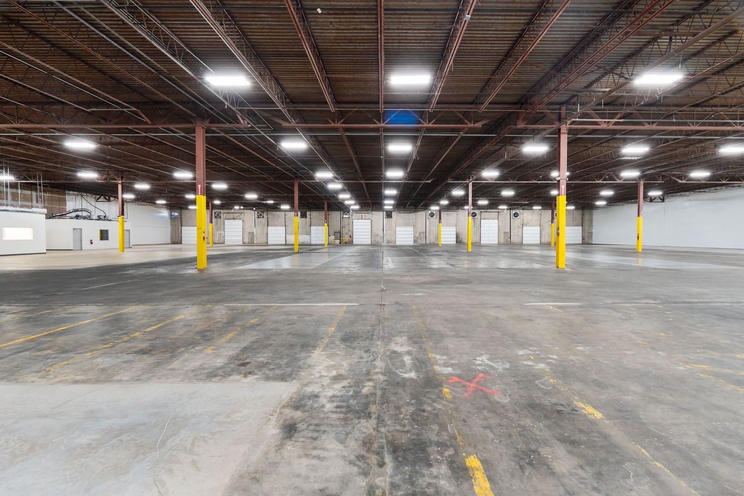 Large empty industrial warehouse interior with concrete floor, support columns, and fluorescent lighting overhead.