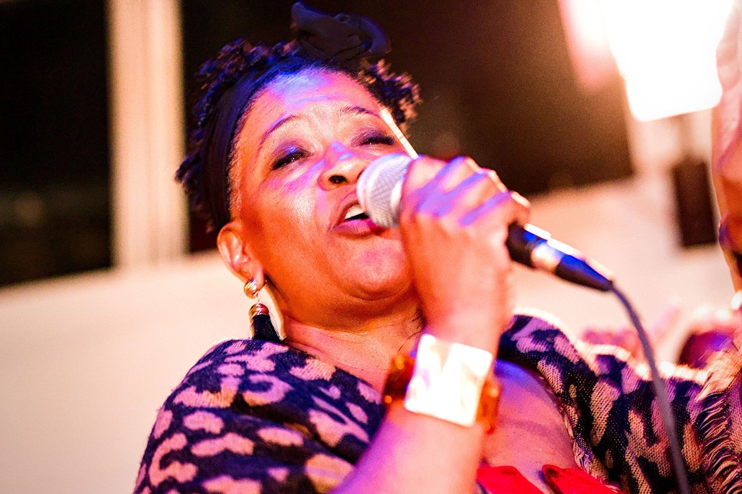 Woman singing passionately into a microphone at an outdoor evening event, wearing a floral top and gold bracelet.