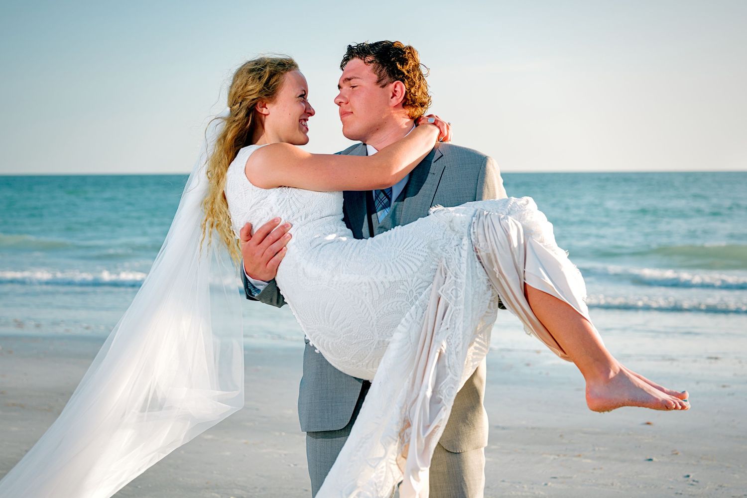 Groom carrying bride in white wedding dress on a sunny beach, both smiling joyfully on their wedding day.