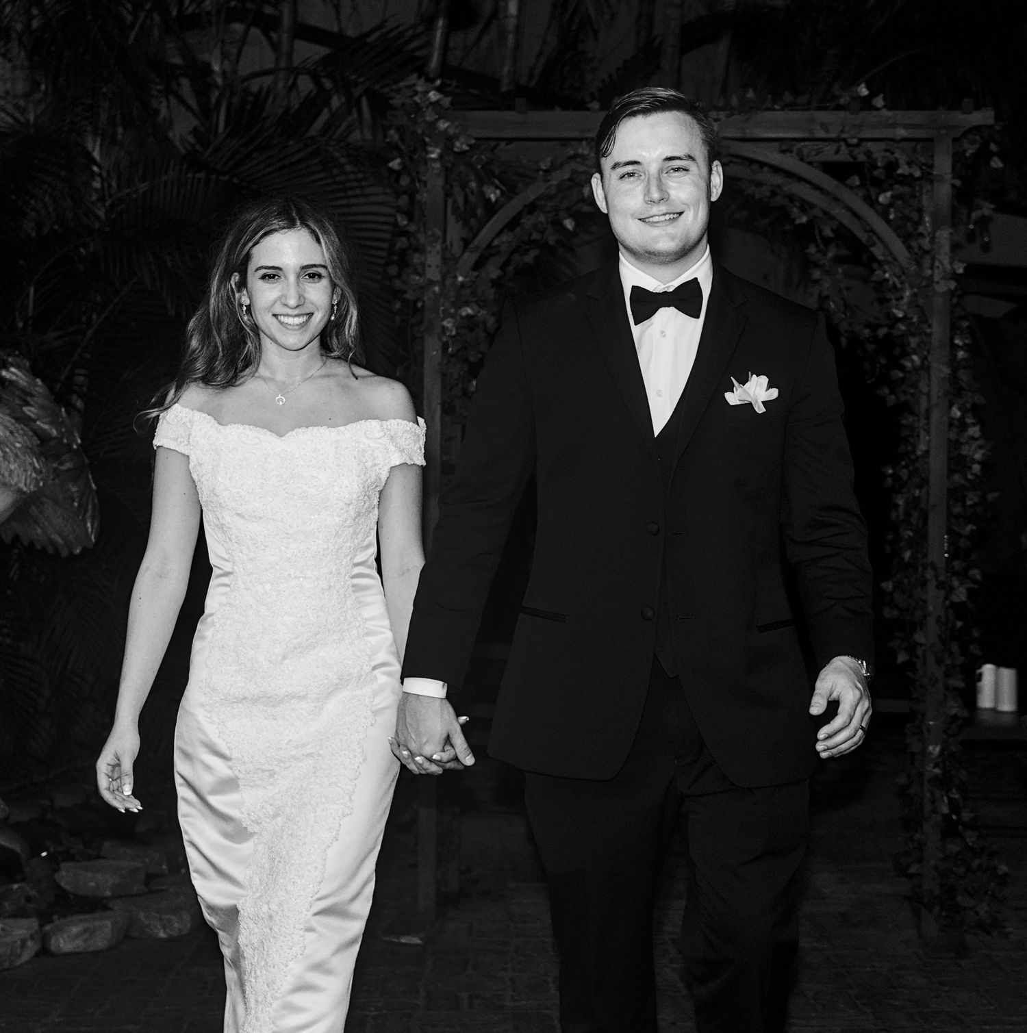 Couple in formal wedding attire holding hands, bride in white off-shoulder gown, groom in black tuxedo, black and white photo.