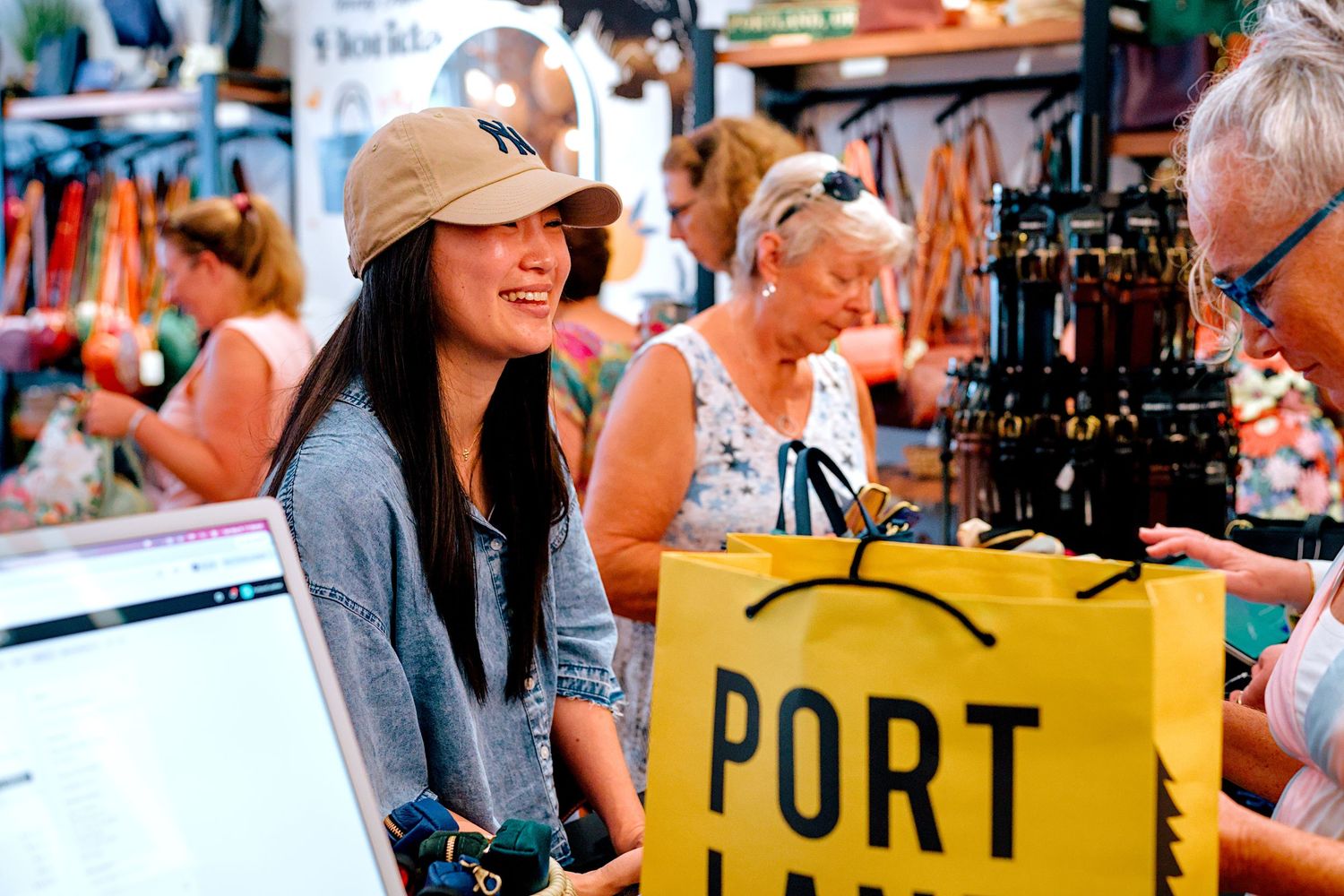 Woman smiling at Portland market stall holding yellow shopping bag, browsing local vendors at busy outdoor bazaar.