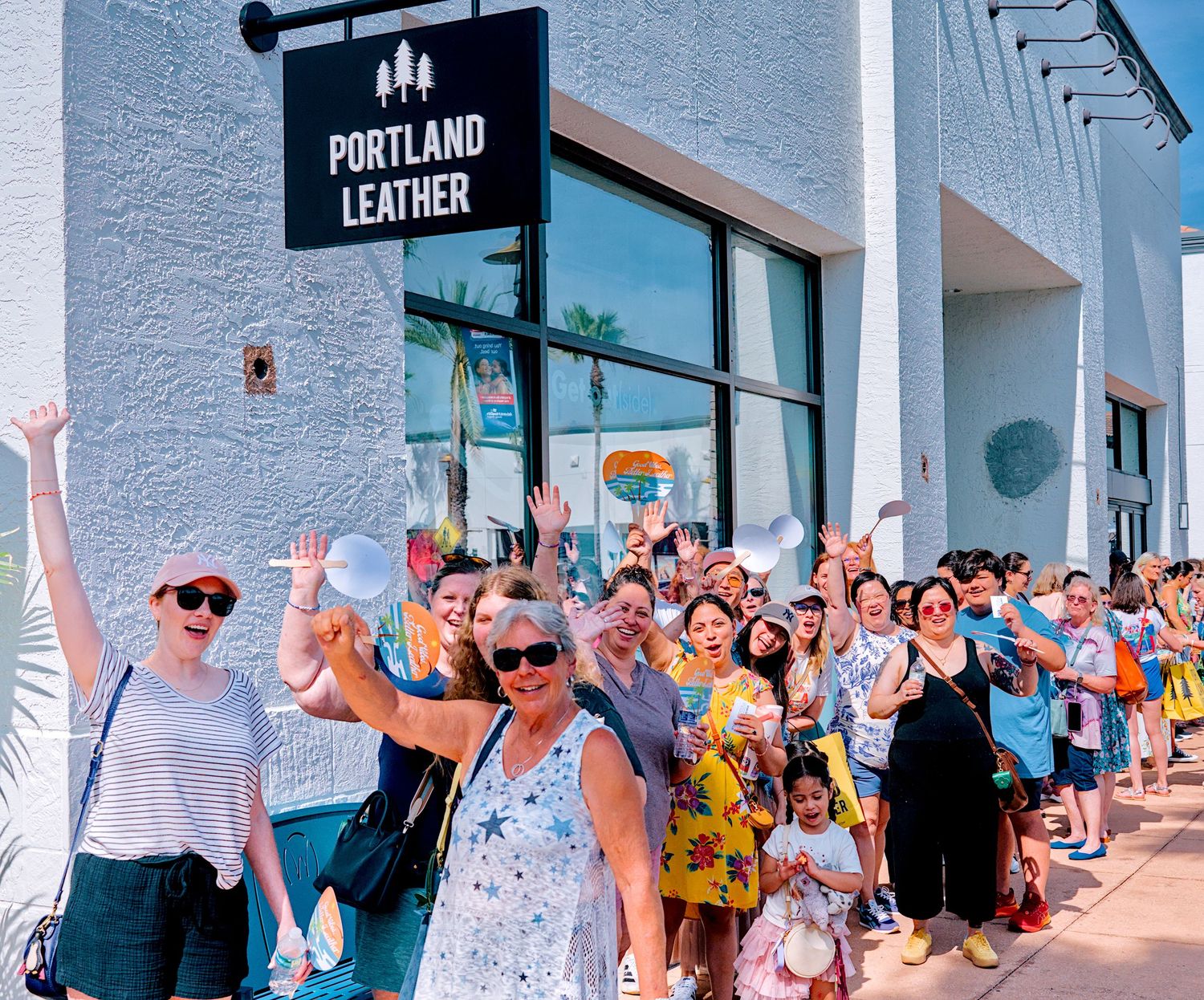 Excited crowd of shoppers gathered outside Portland Leather store on a sunny day, waving and cheering.