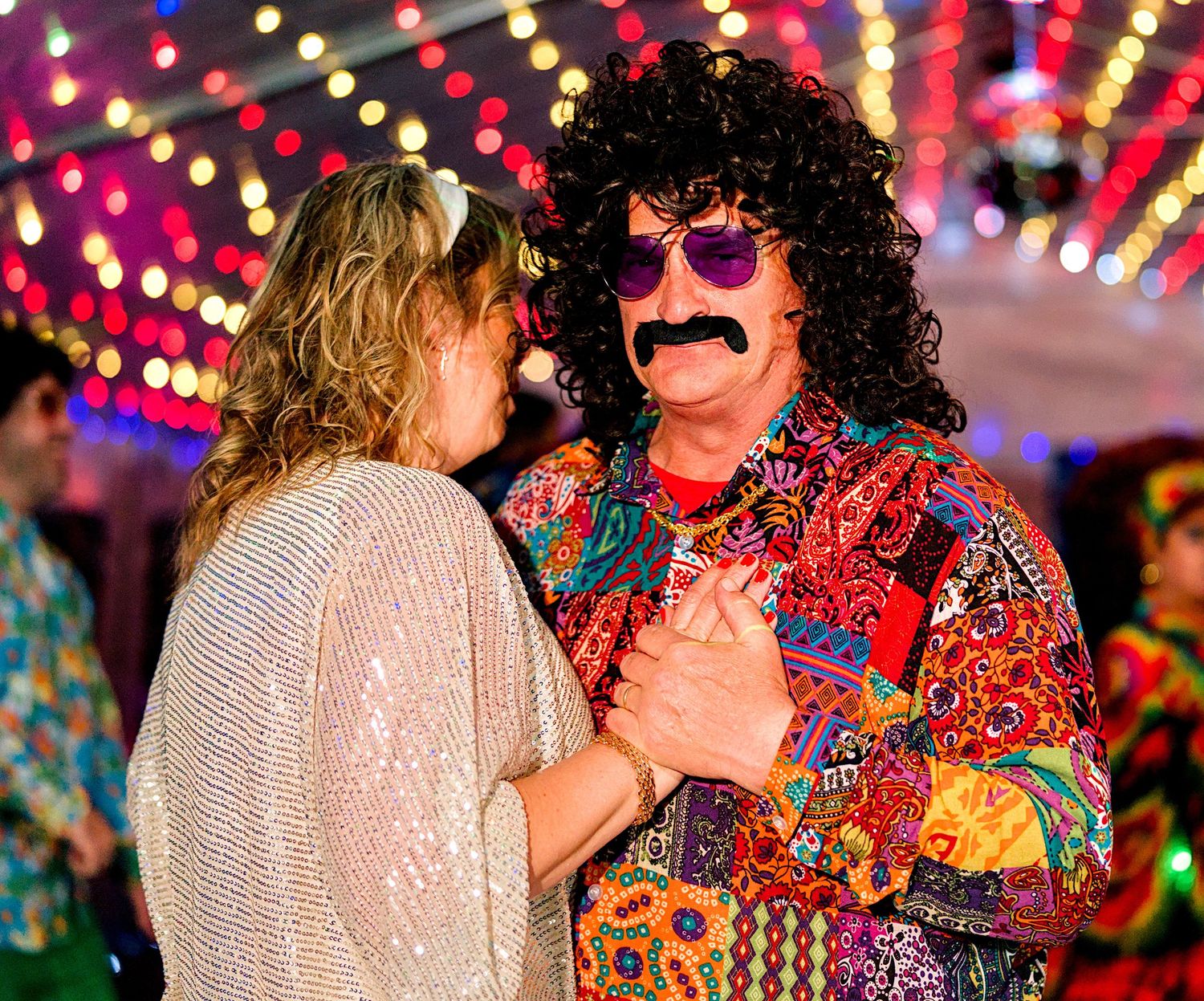 Two party guests in 1970s disco costumes with afro wigs and colorful outfits dancing together at a themed event.