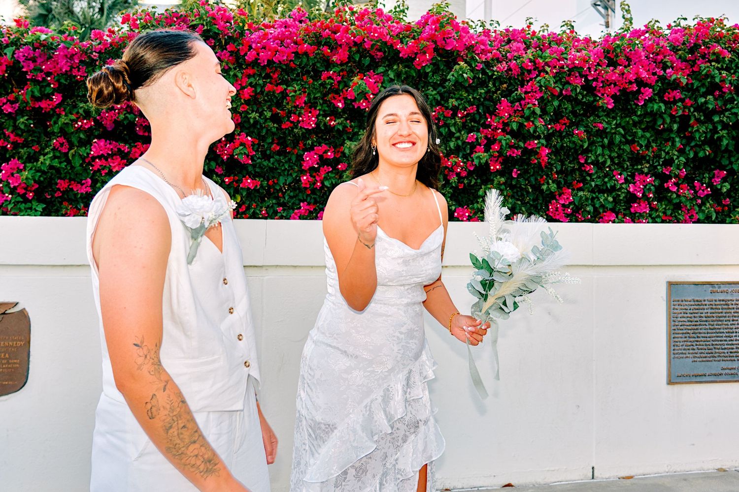 Two brides in white wedding attire laugh together outdoors near vibrant pink bougainvillea wall.