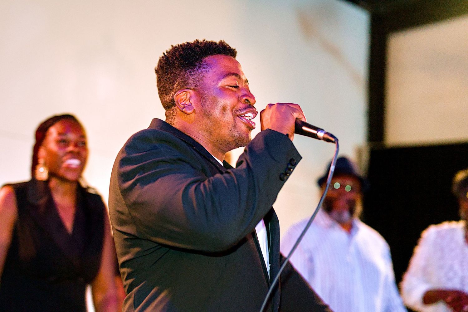 A man in a black suit passionately sings into a microphone on stage at a live event with performers behind him.