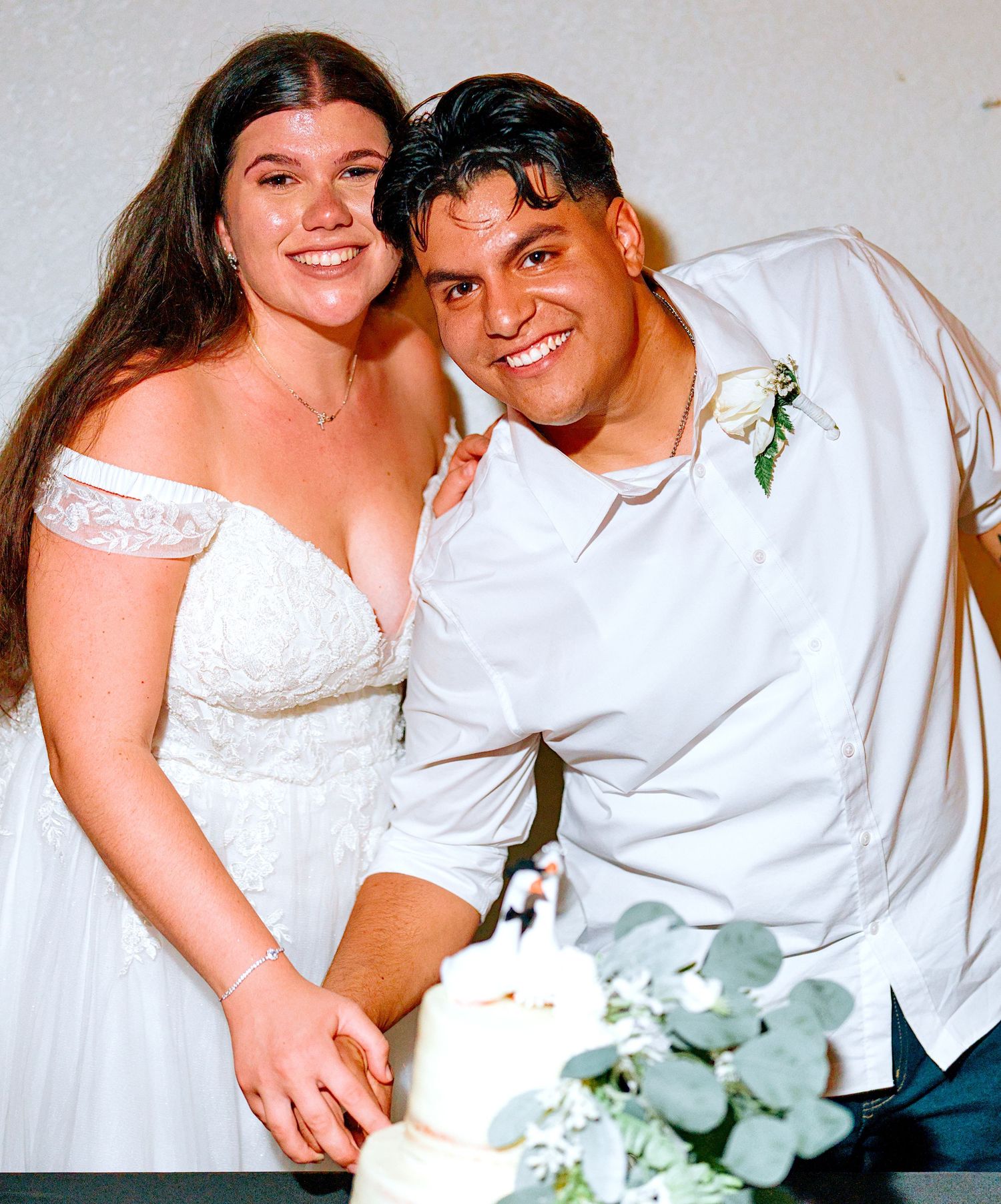 Bride and groom smile while cutting their wedding cake, bride in lace off-shoulder gown, groom in white shirt.
