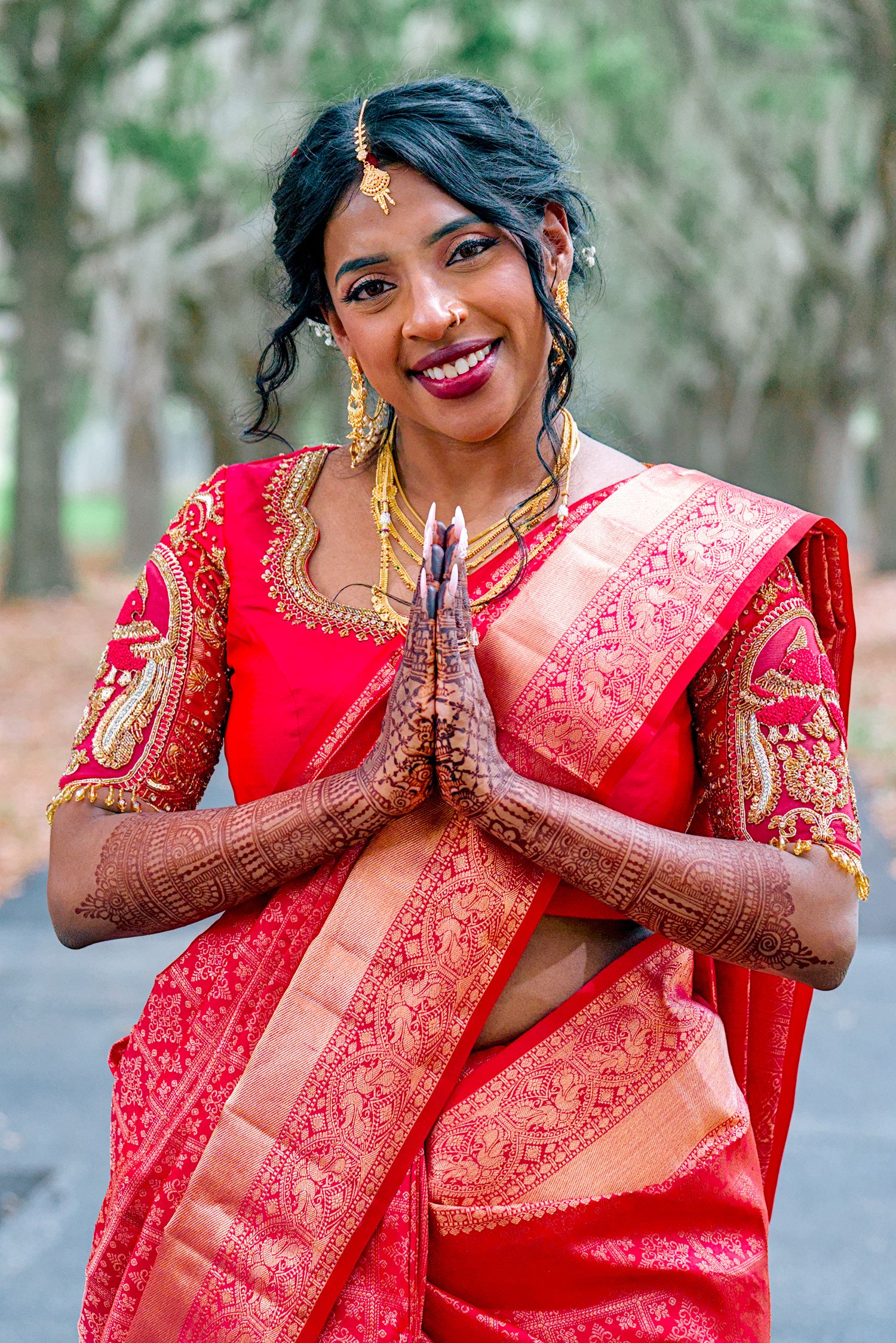 Indian bride in red silk saree with henna hands in namaste pose, wearing gold jewelry and maang tikka.