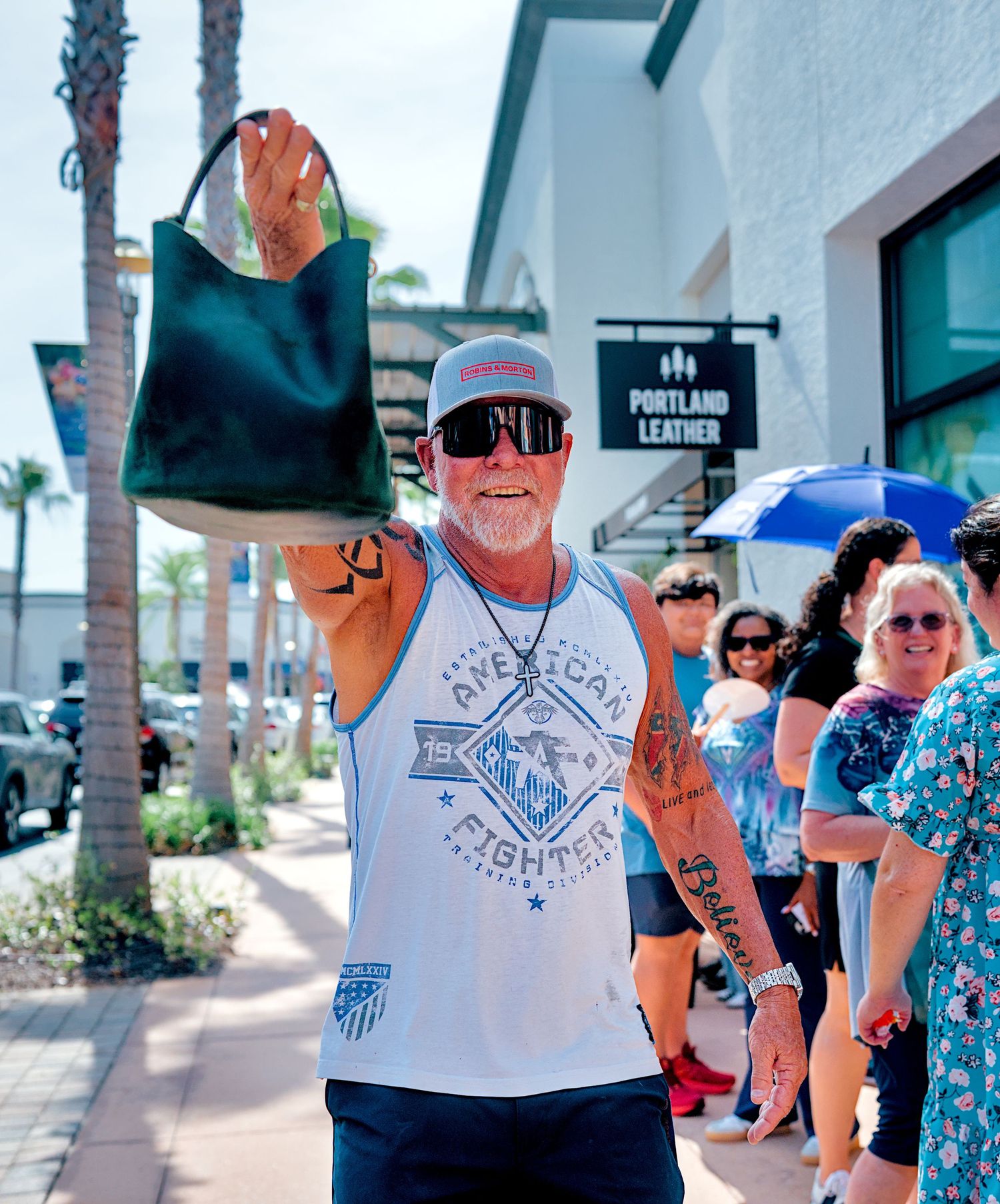 Tattooed man in sleeveless shirt holds up green leather tote bag outside Portland Leather store.