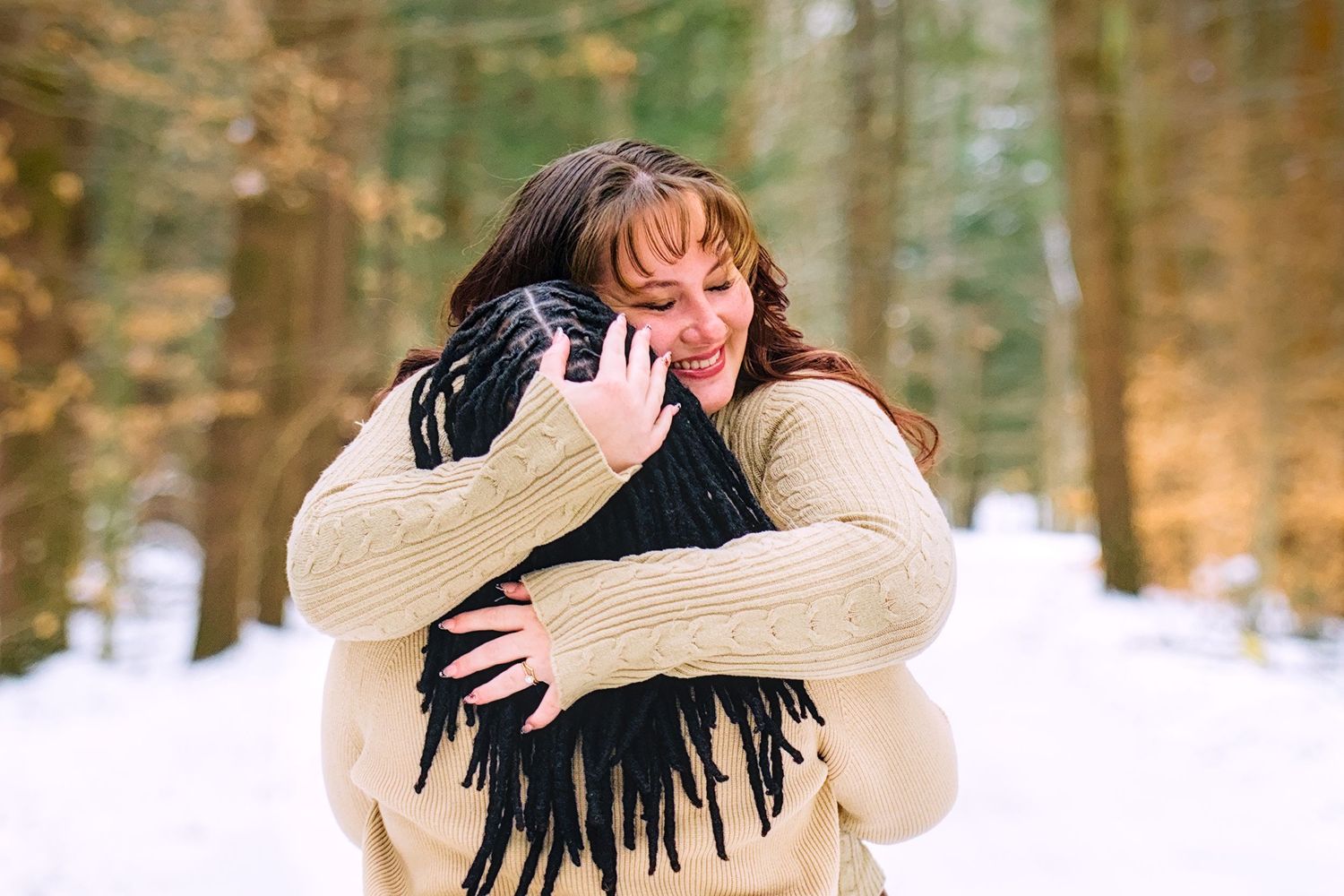 Two women share a warm hug in a snowy winter forest, wearing cozy sweaters and a black fringed scarf.