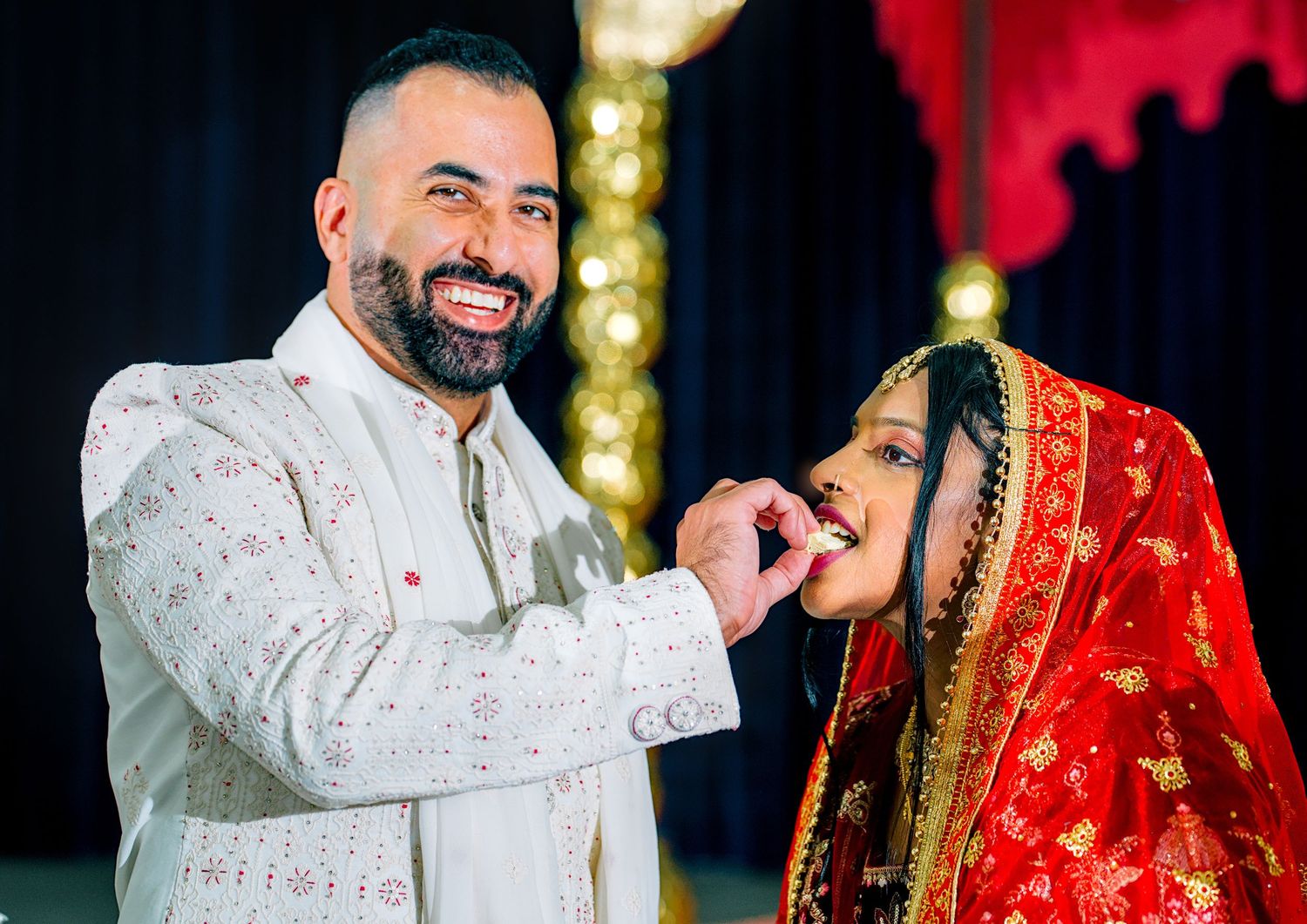 Groom in white sherwani feeding bride in red saree during Indian wedding ceremony.