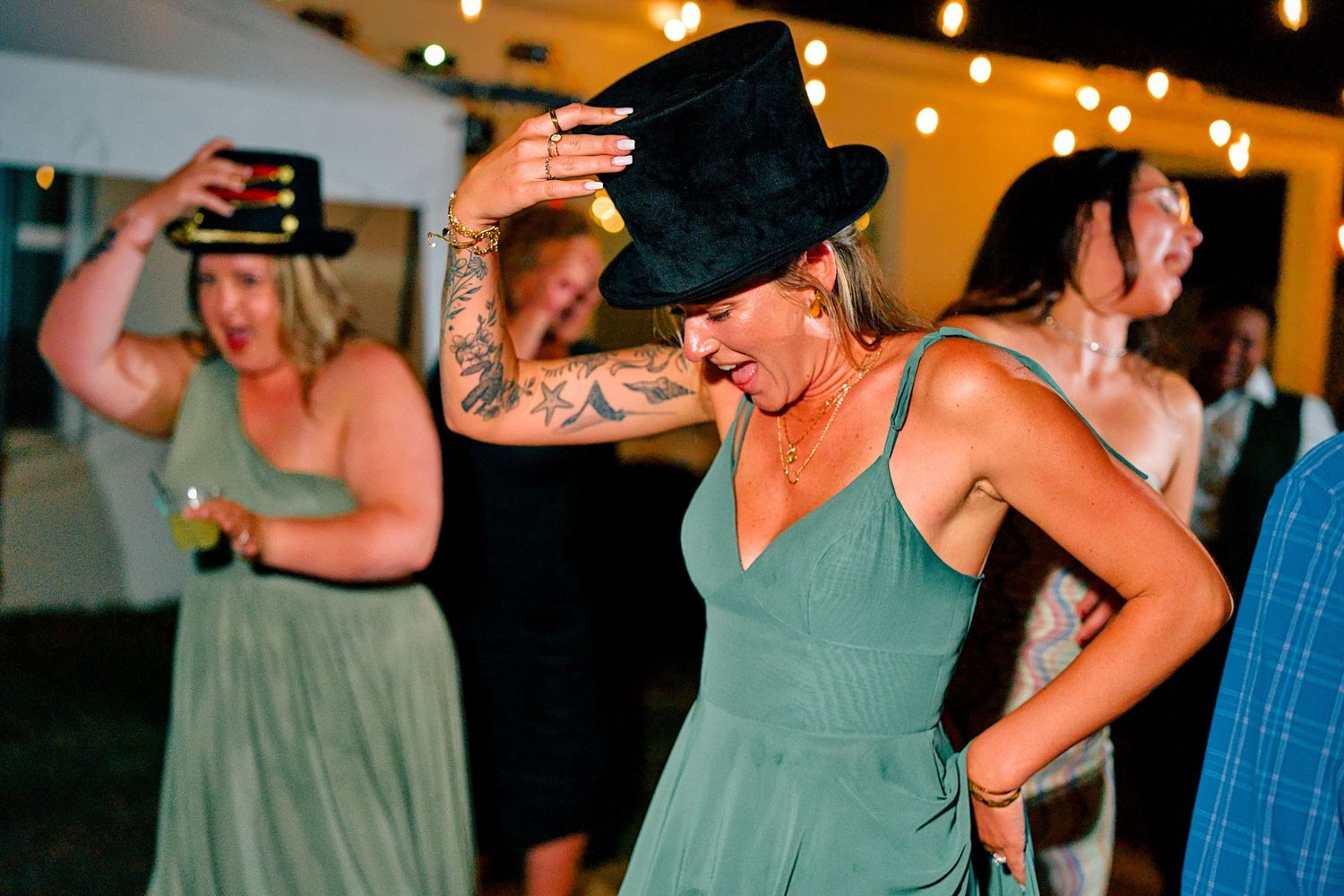 Bridesmaid in green dress dancing with top hat at lively wedding reception celebration.
