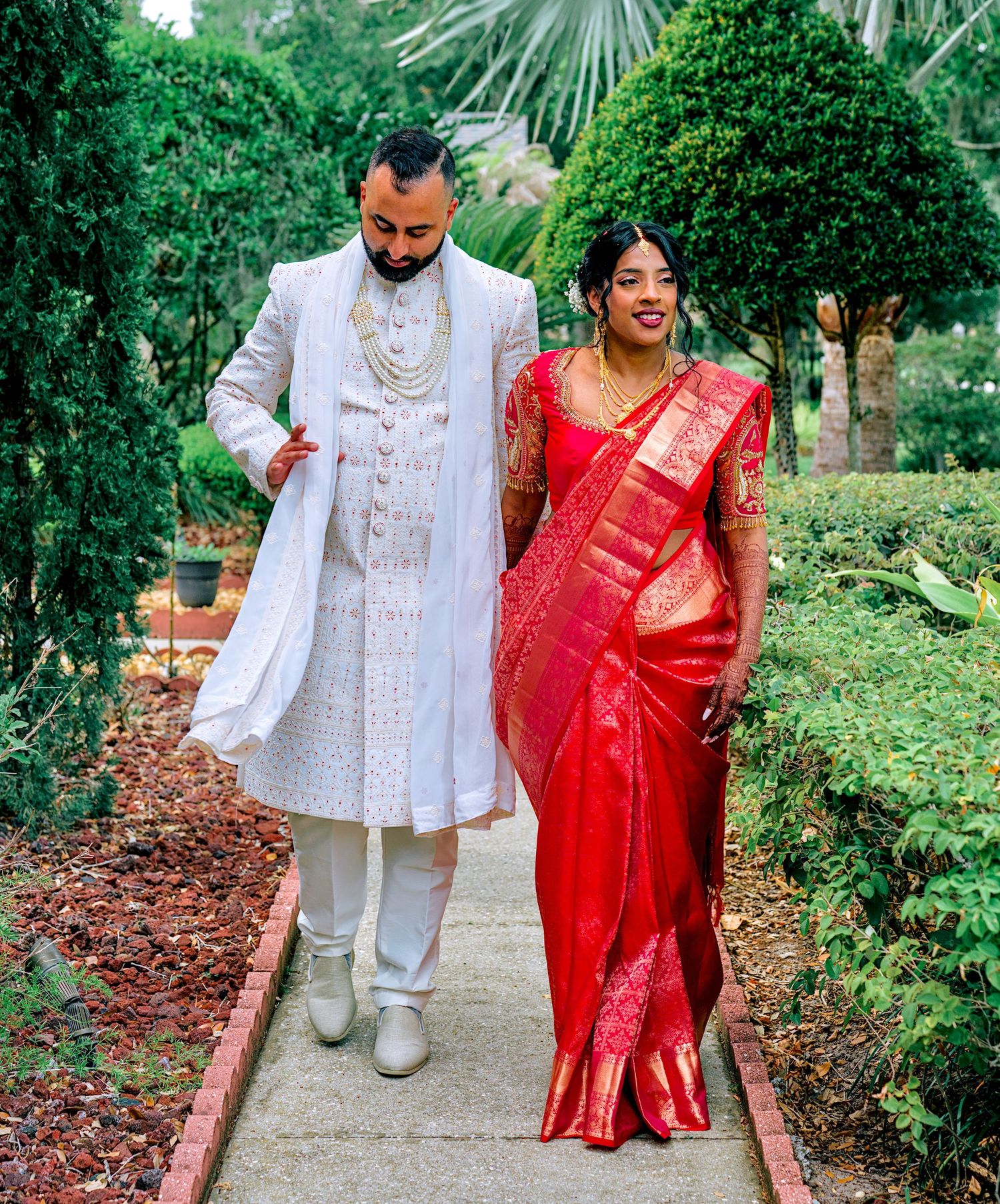 Indian couple in traditional wedding attire walk garden path, groom in white sherwani, bride in red silk saree.
