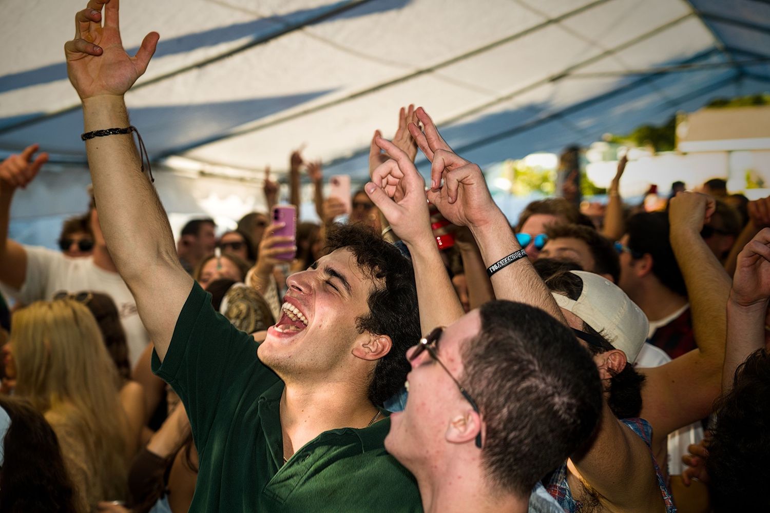 A crowd of excited spectators with raised arms cheering enthusiastically under a white tent at an outdoor event.