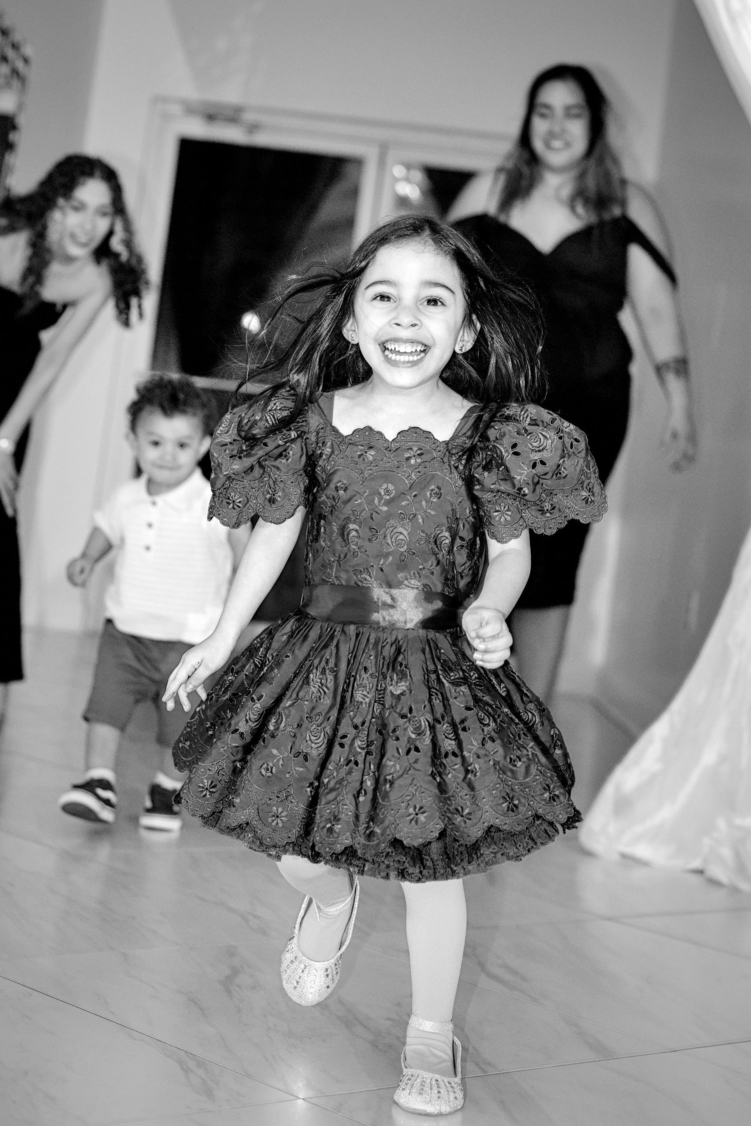 A happy young girl in a black lace dress dances joyfully at a formal event with guests in background.