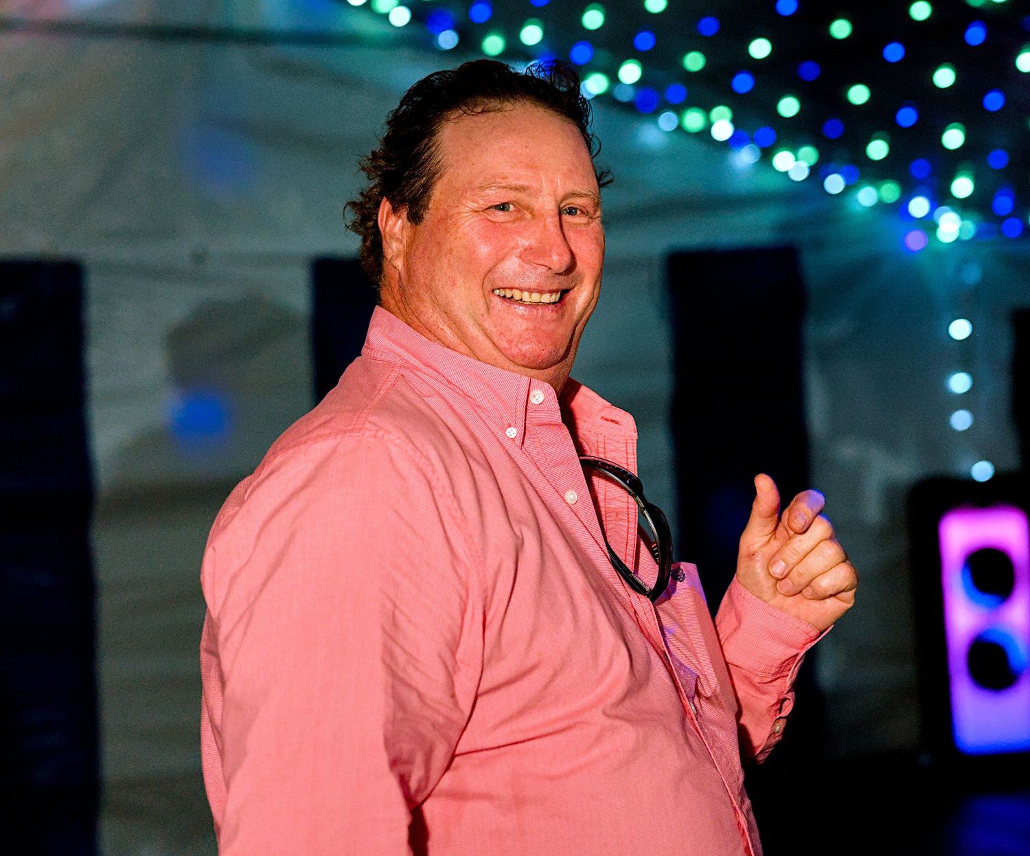 Smiling man in pink shirt with stethoscope giving thumbs up at colorful event with bokeh lights.