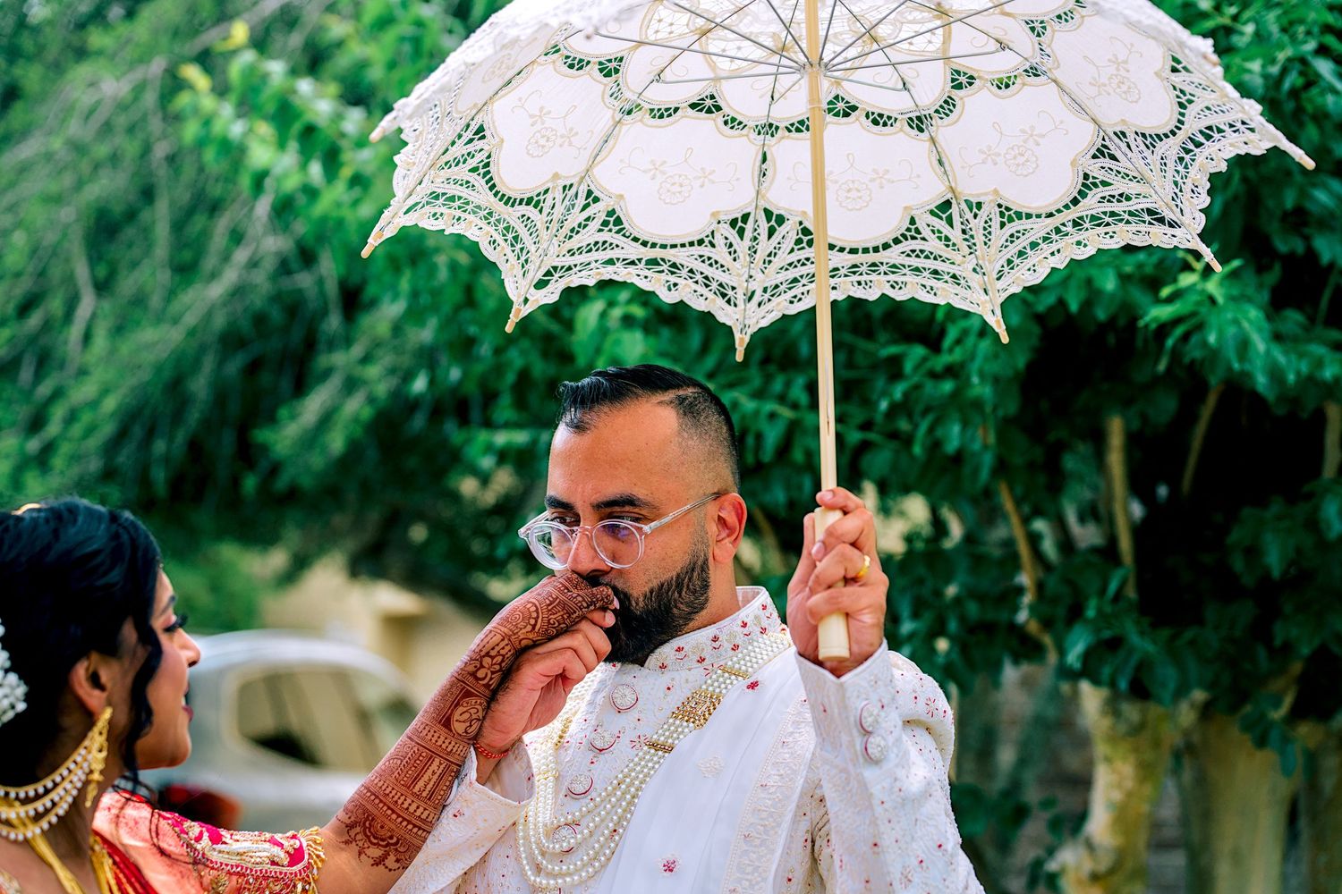 Groom in white sherwani holding lace parasol during outdoor Hindu wedding.