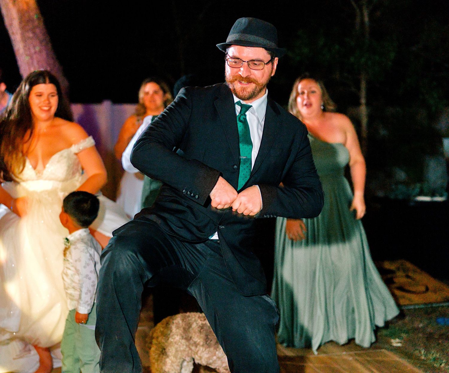 Man in suit and fedora dancing energetically at a wedding reception with bridesmaids in sage green dresses in background.