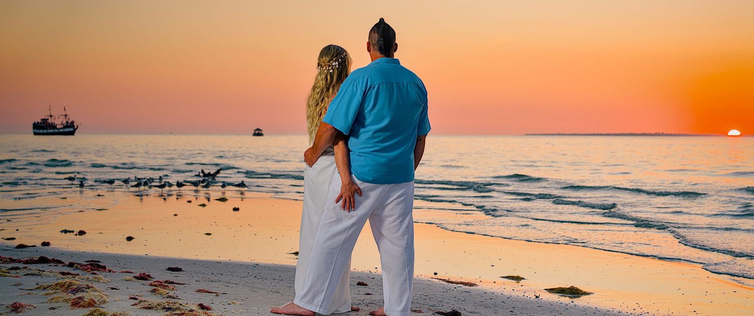 Romantic couple embracing on a sandy beach watching a stunning orange and pink sunset over calm ocean waters.