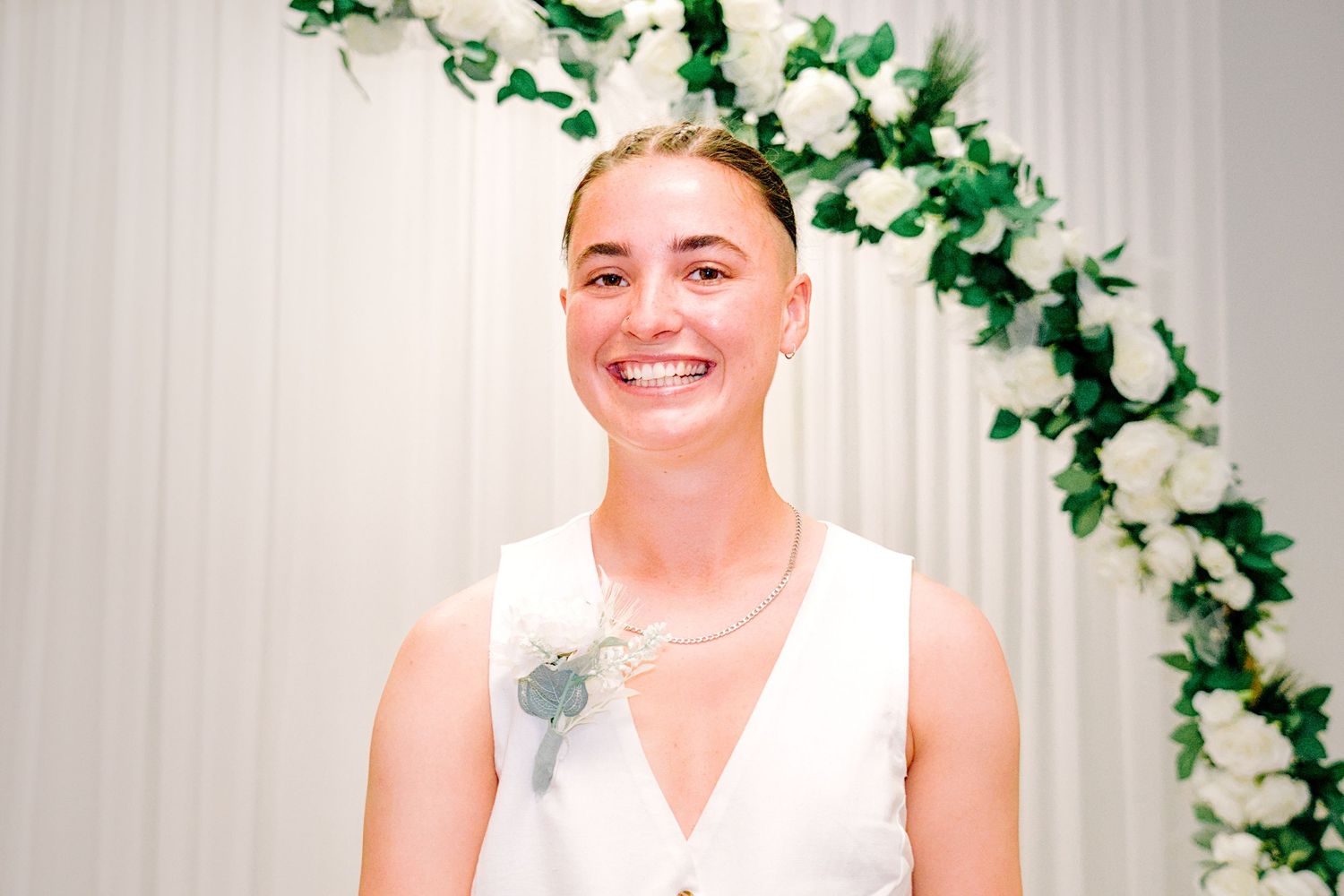 Smiling bride in white dress stands before floral arch with white roses and greenery at wedding ceremony.