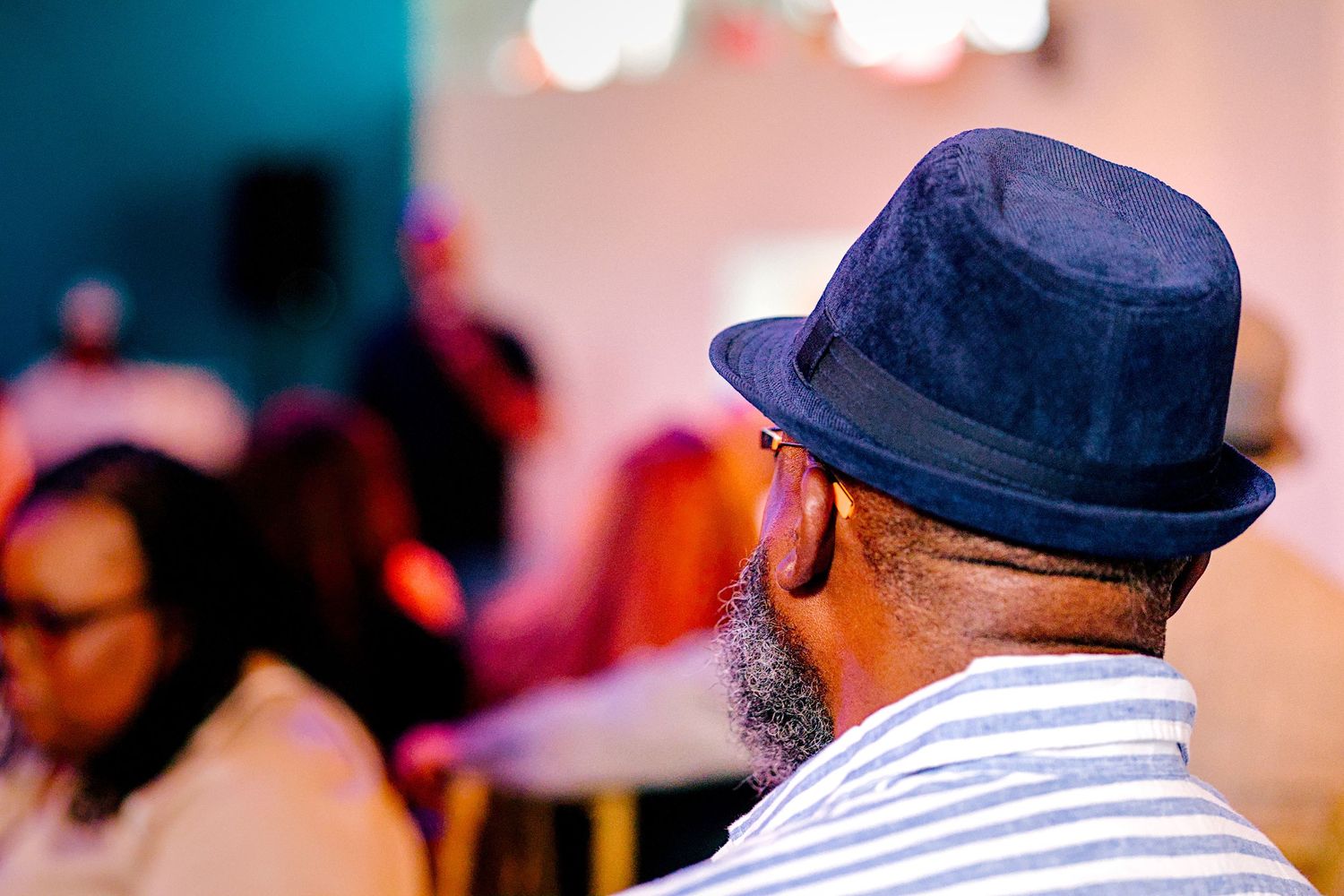 Man wearing a navy blue fedora hat at a social gathering, viewed from behind in a striped shirt.