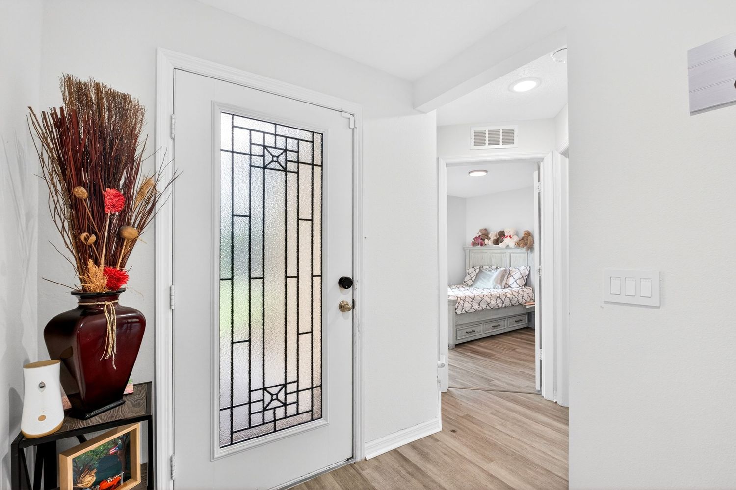 Bright entryway with decorative glass front door, hardwood floors, and a hallway leading to a bedroom.