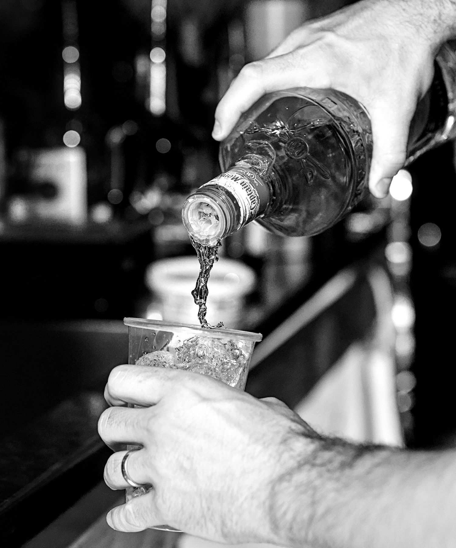 Black and white photo of a bartender pouring liquor from a bottle into an ice-filled cup.