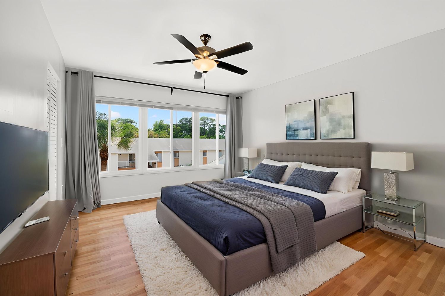 Bright bedroom with gray upholstered bed, hardwood floors, ceiling fan, and large windows allowing natural light.