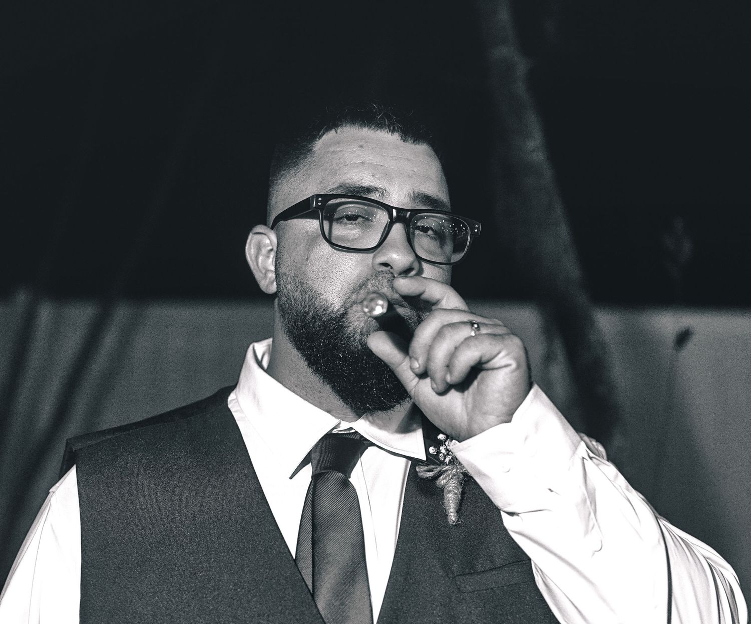 Bearded man in vest and tie smokes cigar at formal event in black and white photo.