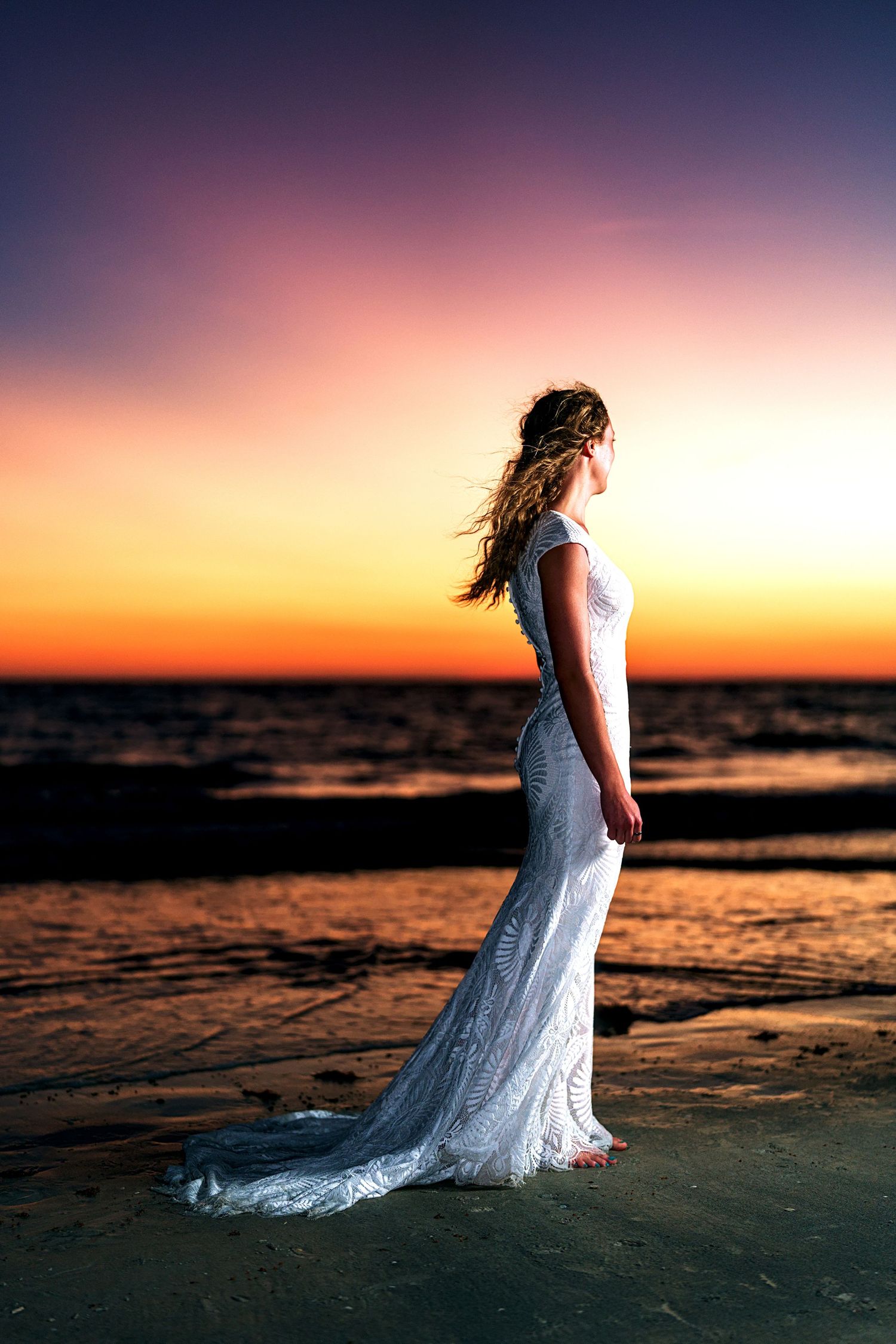 Bride in white lace gown standing on beach at sunset with vibrant orange and purple sky.
