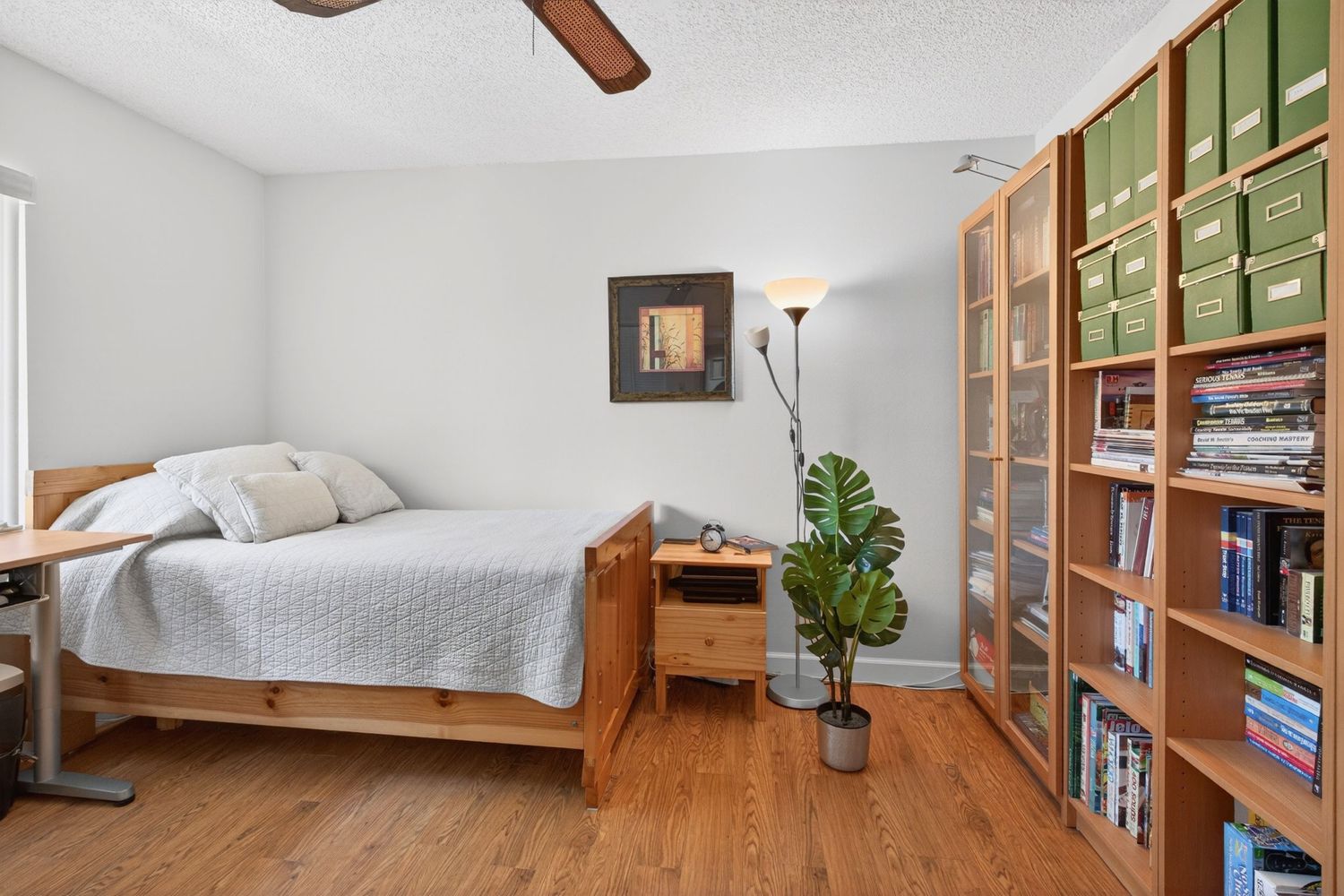 Cozy bedroom with wood-frame bed, hardwood floors, ceiling fan, and tall bookshelf filled with books and storage boxes.