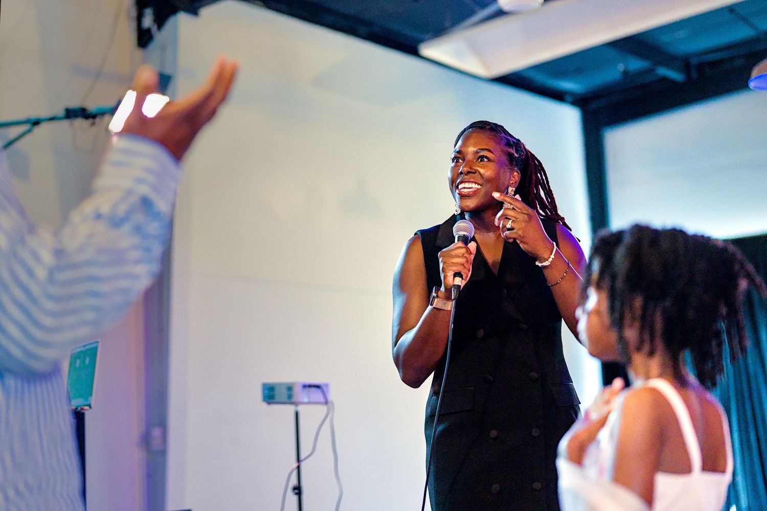 A woman speaks into a microphone on stage, smiling confidently in a black dress at an indoor event.