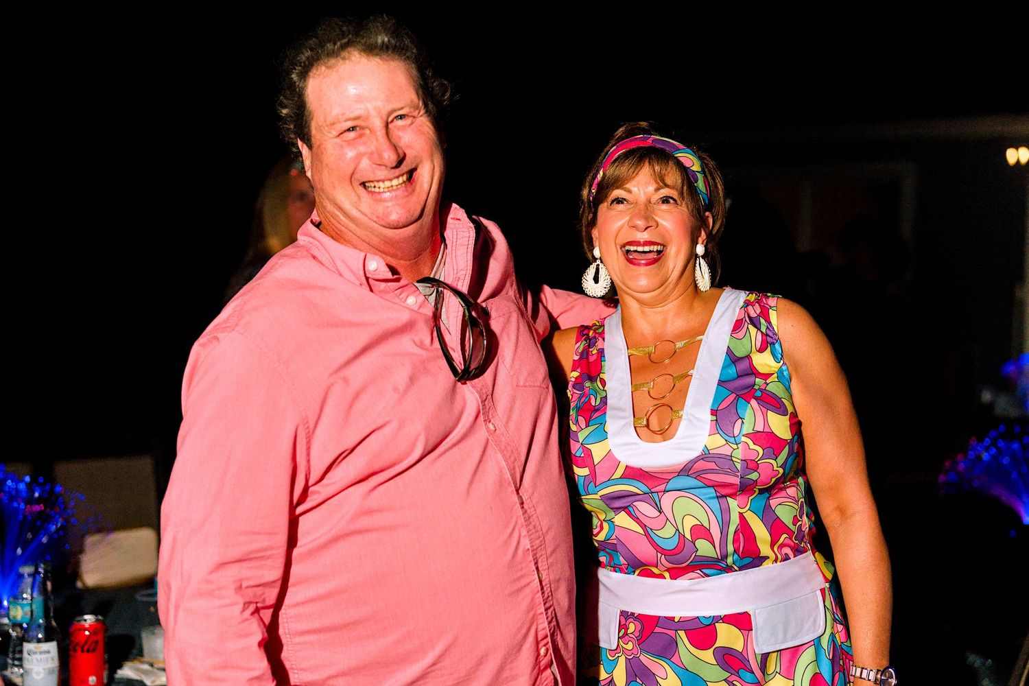 A smiling man in a pink shirt and a woman in a colorful dress pose together at an outdoor evening event.
