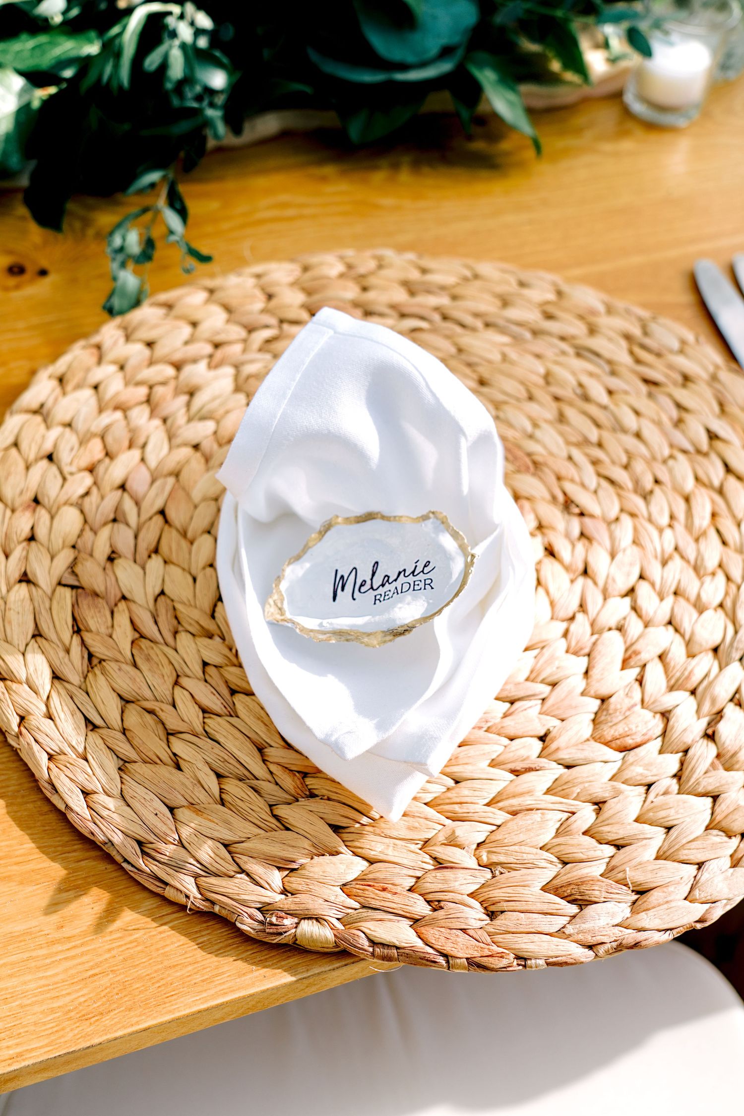 Elegant place setting with woven charger, white napkin, and oyster shell name card reading Melanie at a wedding reception.