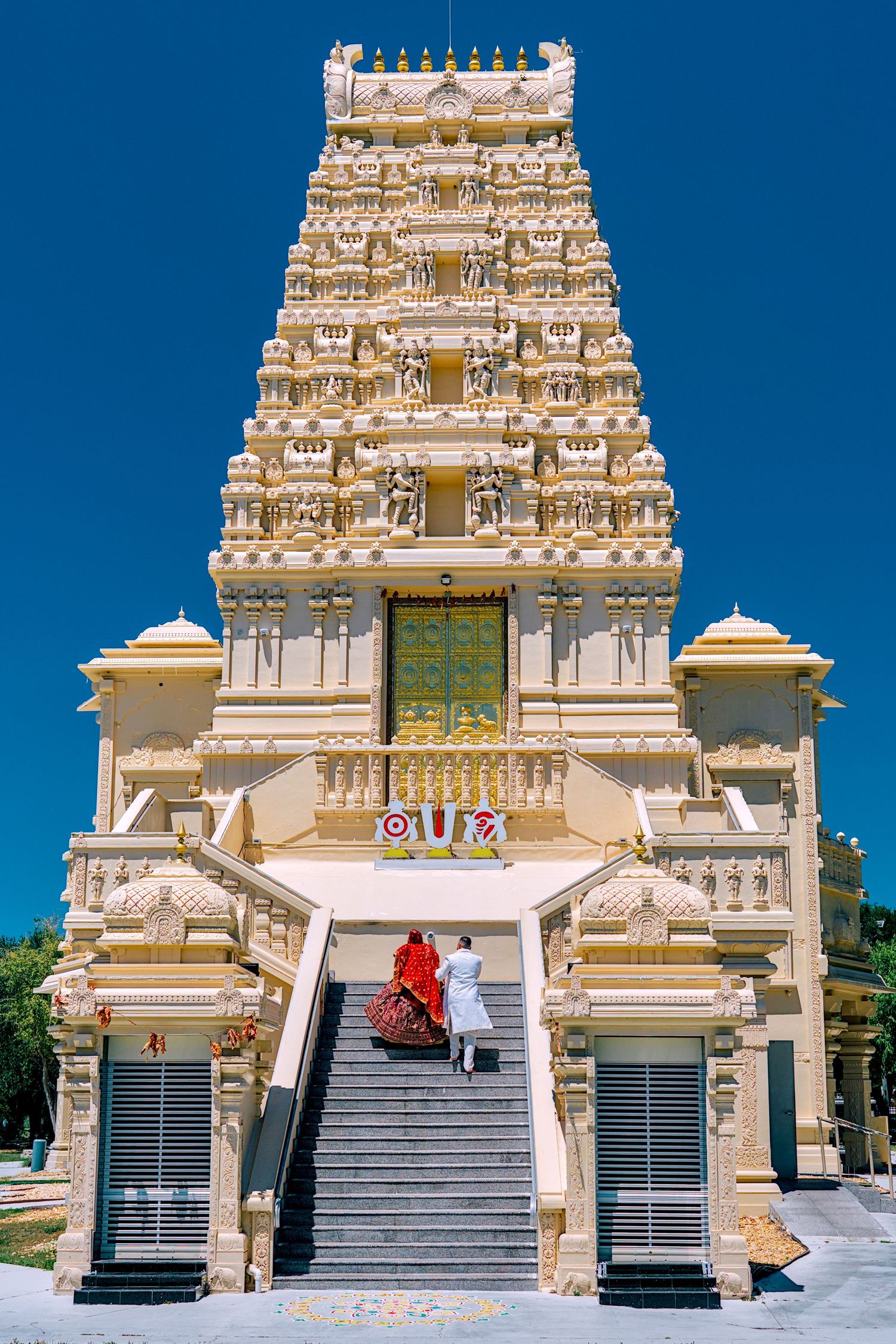 Couple in traditional Indian wedding attire climbing stairs of ornate Hindu temple gopuram.