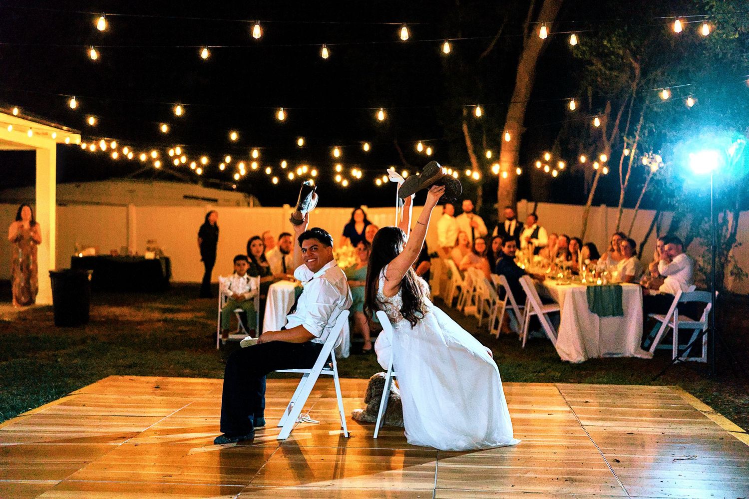 Bride and groom share a romantic first dance on an outdoor wooden dance floor under warm string lights at night.
