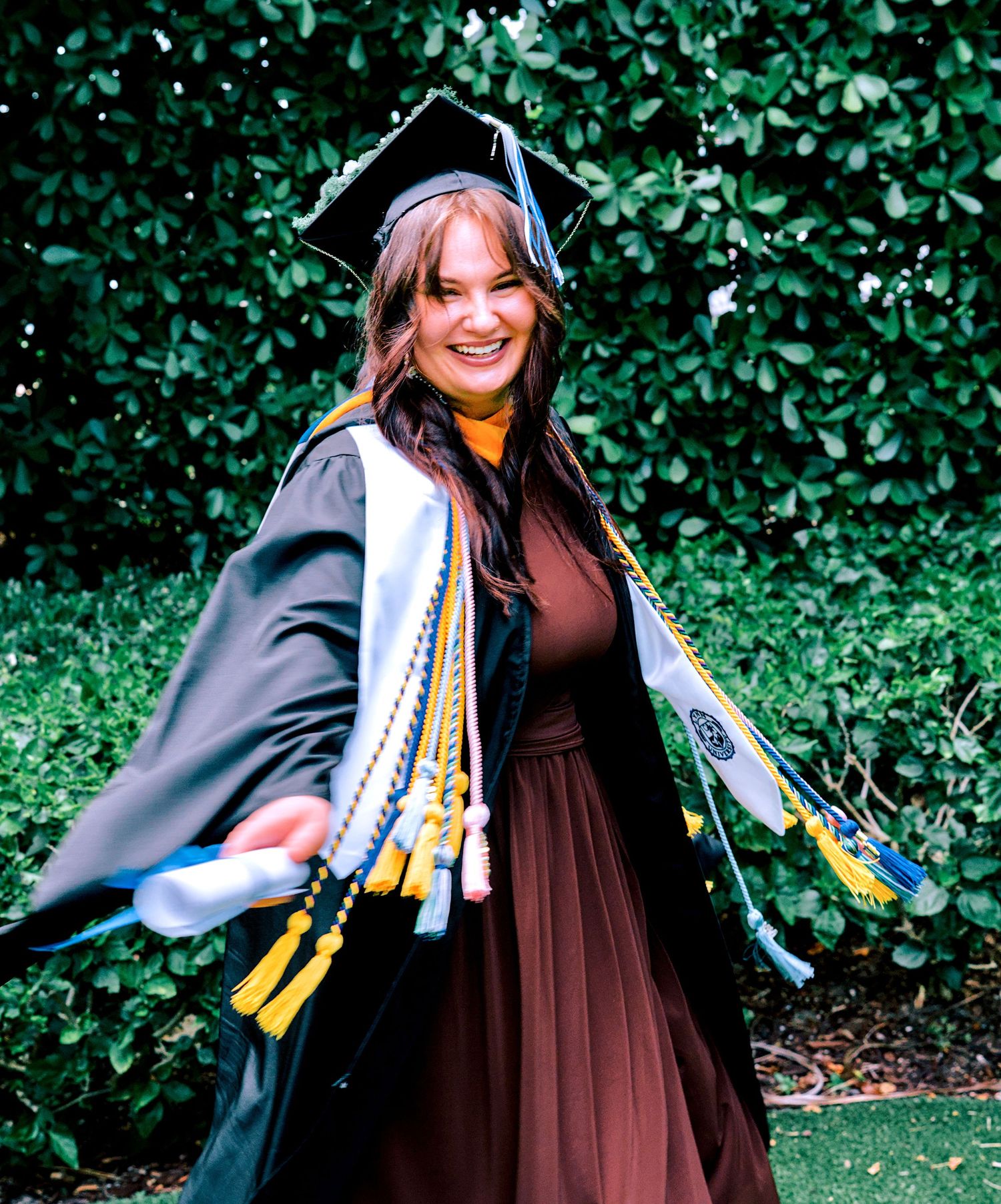 Joyful graduate in black cap and gown with colorful honor cords outdoors.
