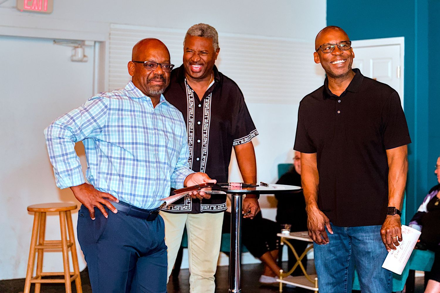 Three smiling men posing together indoors near a bar stool in a bright modern space.