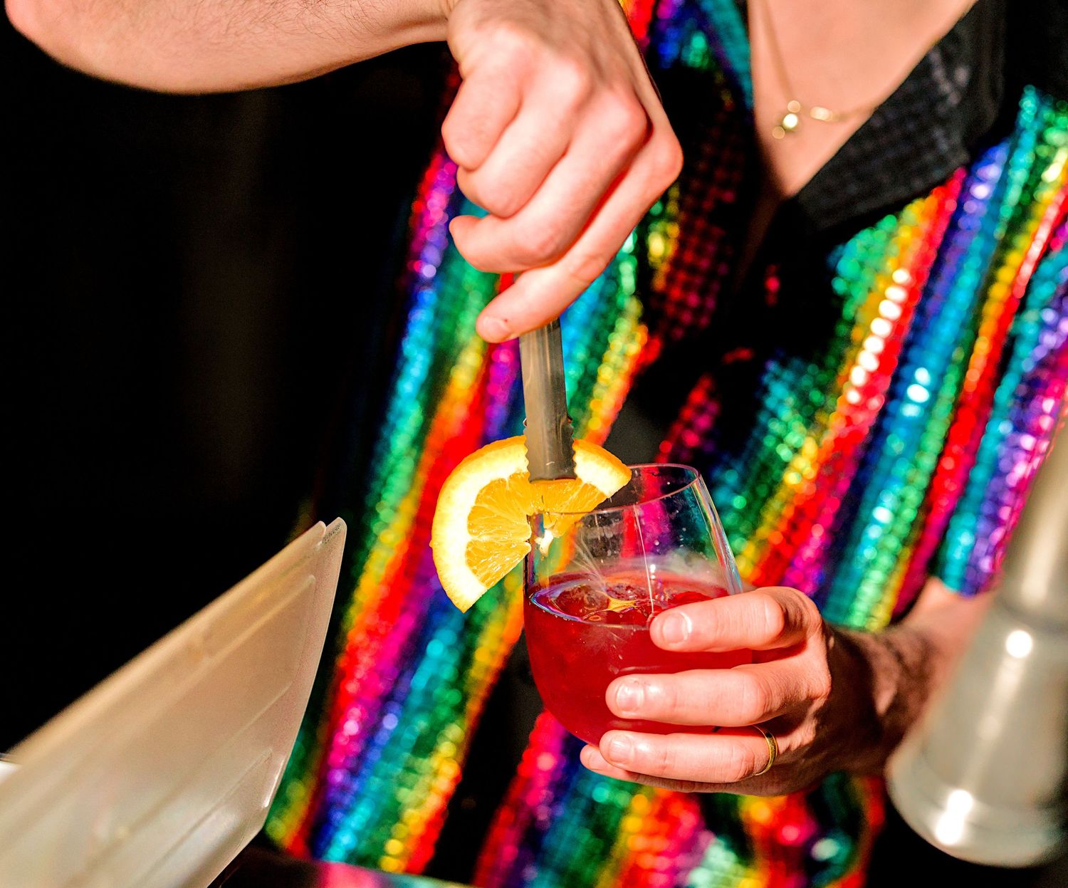Bartender in rainbow sequin jacket garnishing a red cocktail with lemon slice and straw.