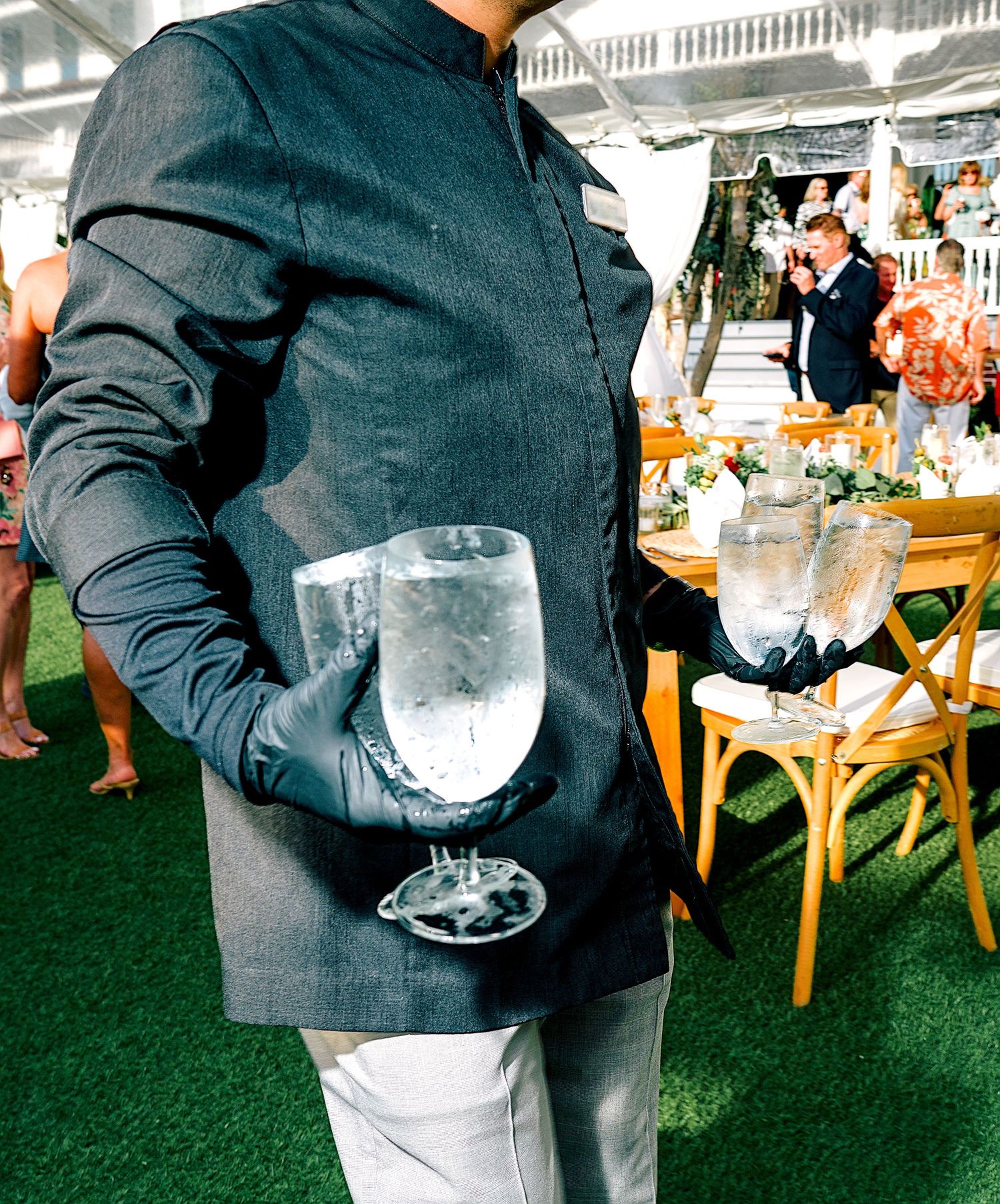 A waiter in a dark mandarin collar jacket holds a large water goblet at an elegant outdoor wedding reception.