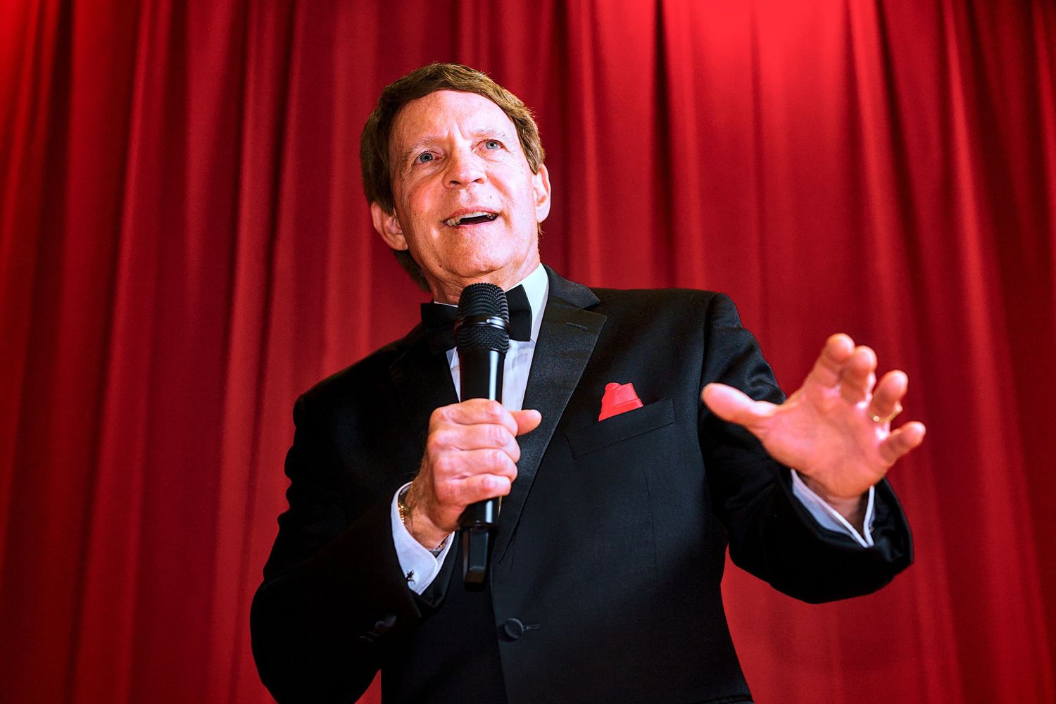 Performer in black tuxedo gesturing dramatically against red curtain backdrop.