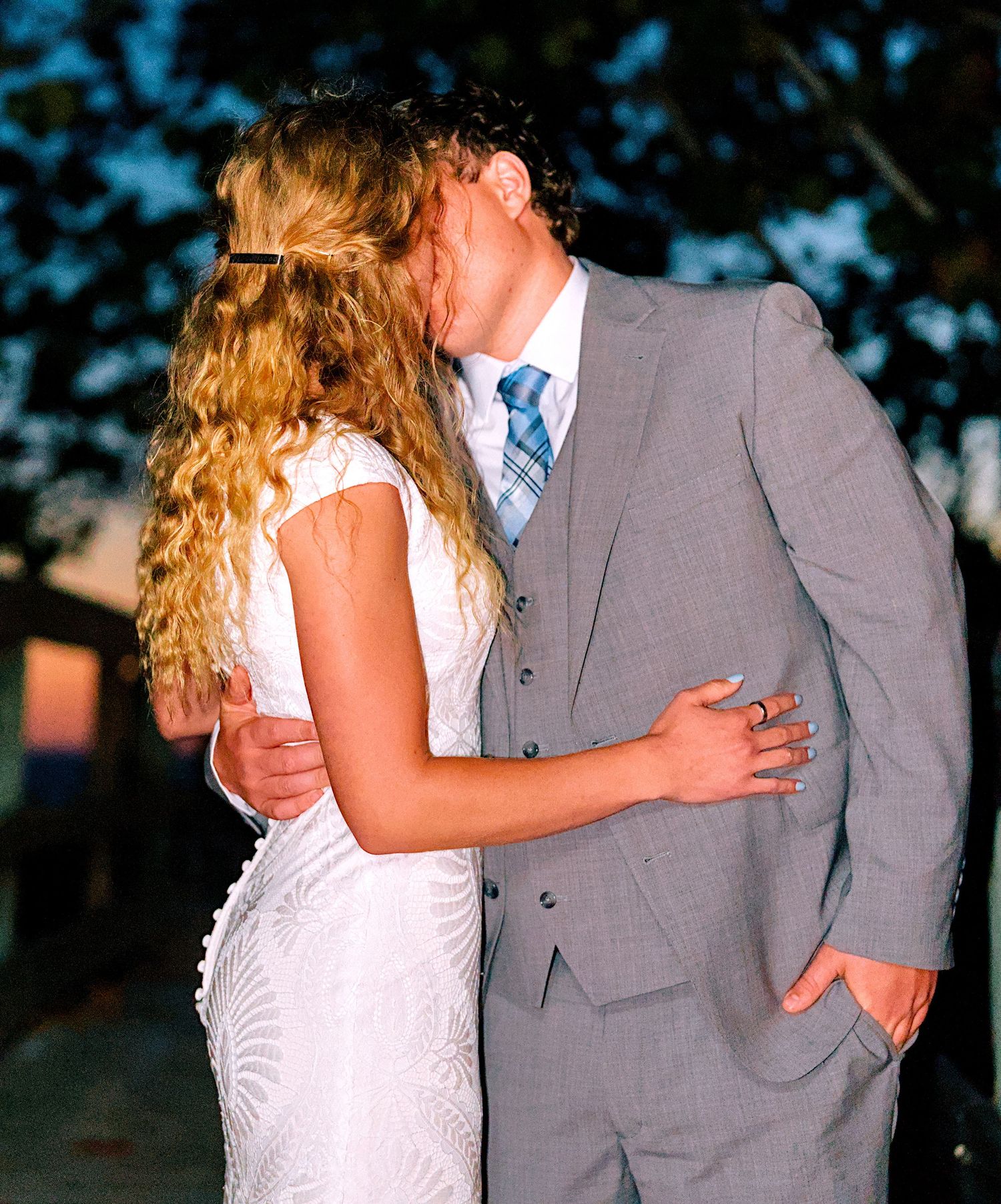 A bride and groom share a romantic kiss outdoors, with the bride in a white lace gown and groom in a gray suit.