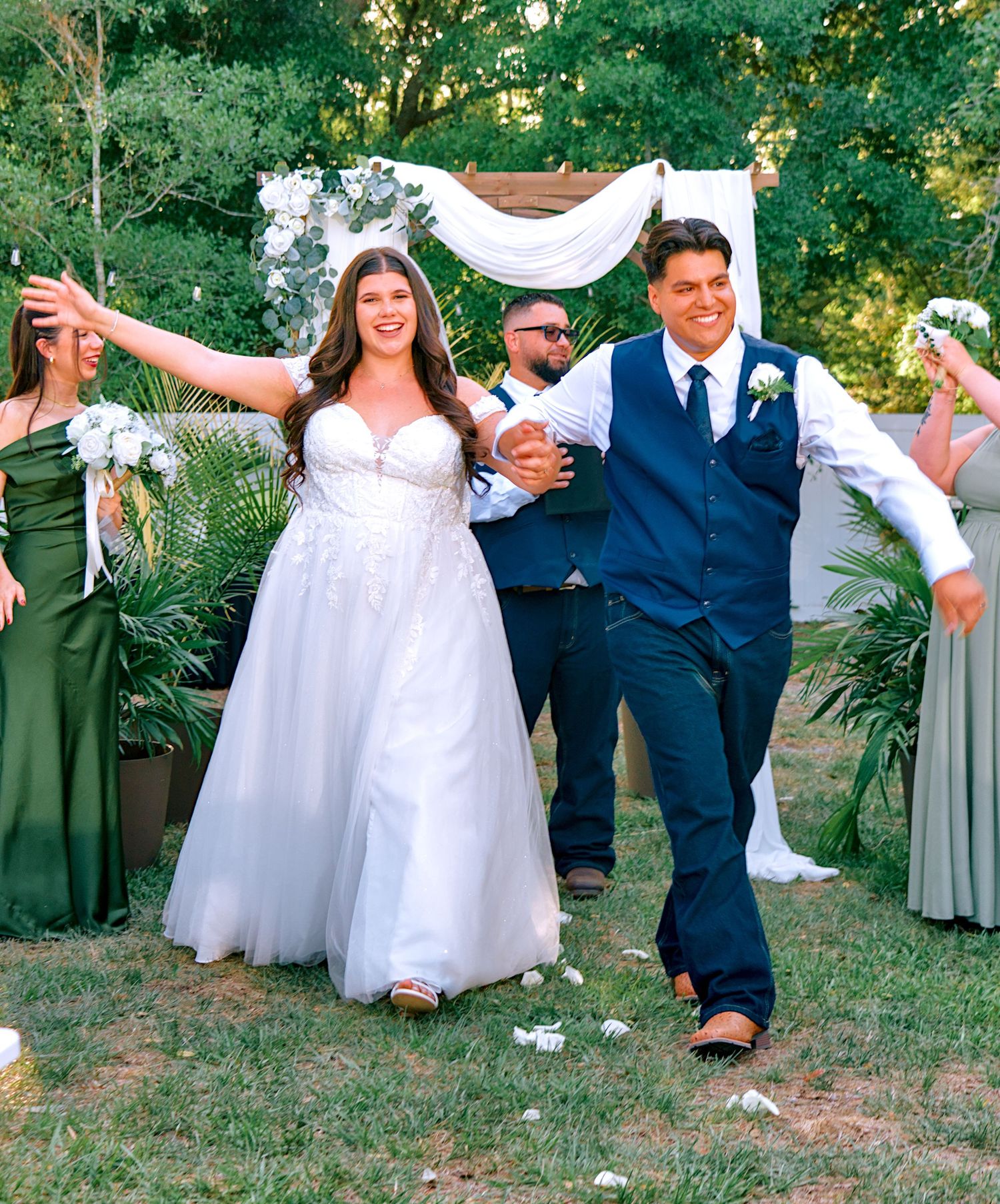 Joyful bride and groom walk down outdoor wedding aisle, surrounded by bridesmaids in green dresses and floral arch backdrop.