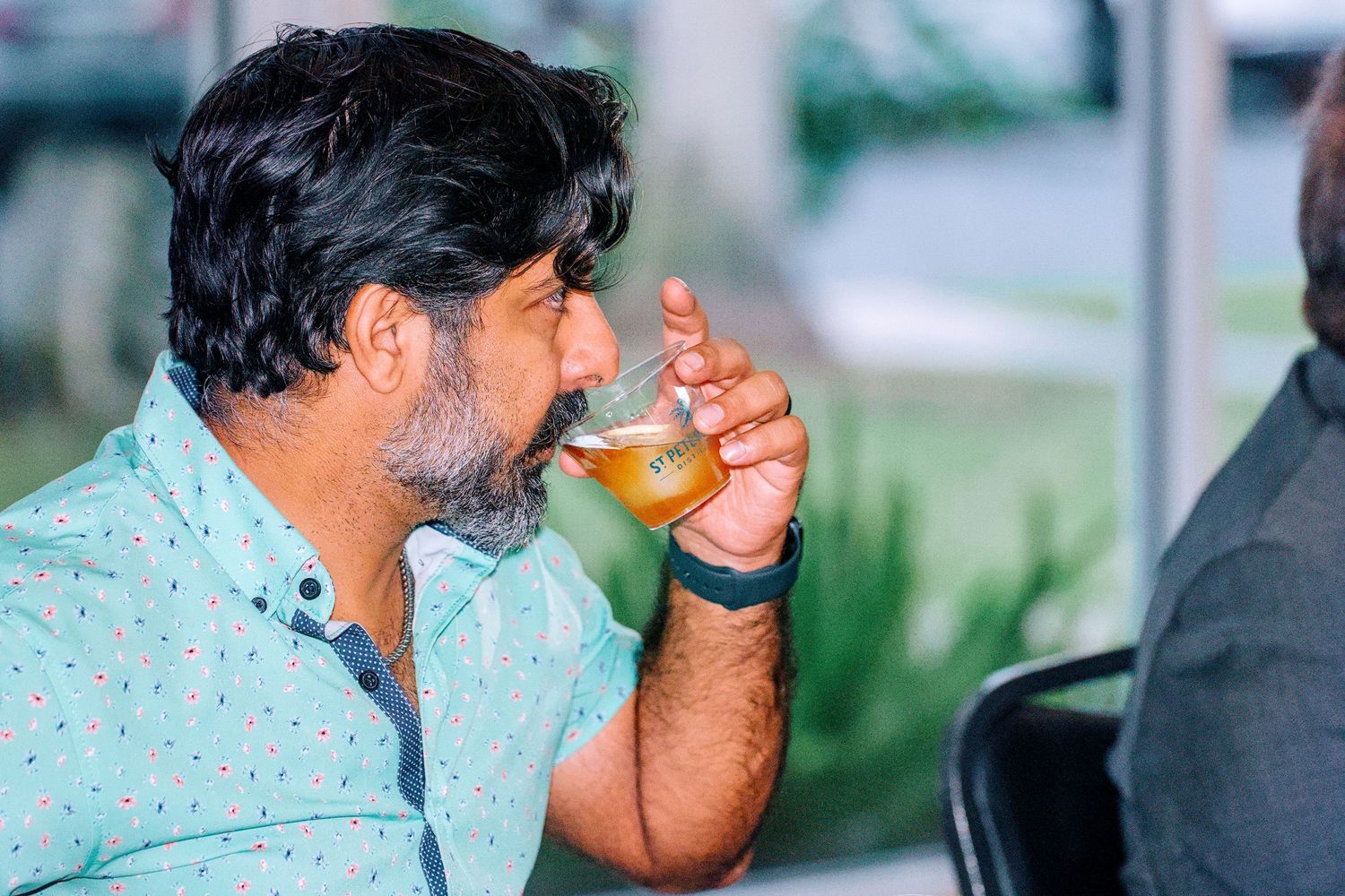 A bearded man in a teal shirt sipping a glass of craft beer outdoors at a social gathering.