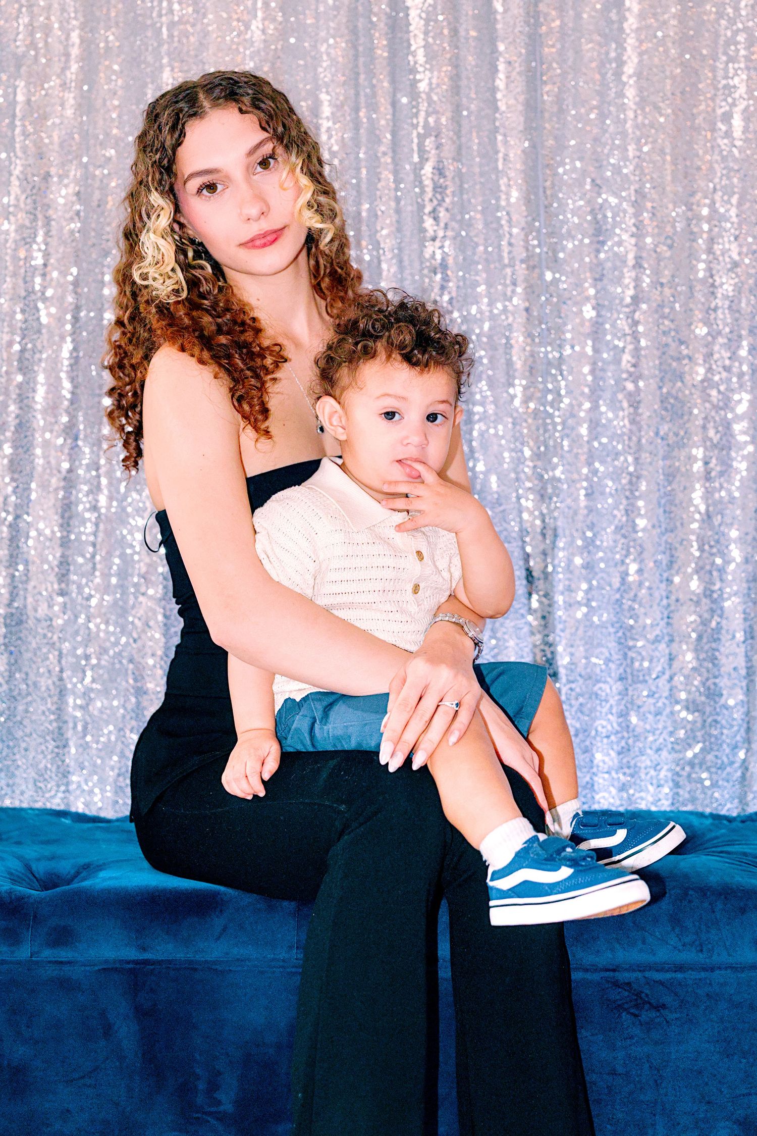 Young woman with curly hair holds a toddler in sneakers, posing on blue velvet bench before sequin backdrop.