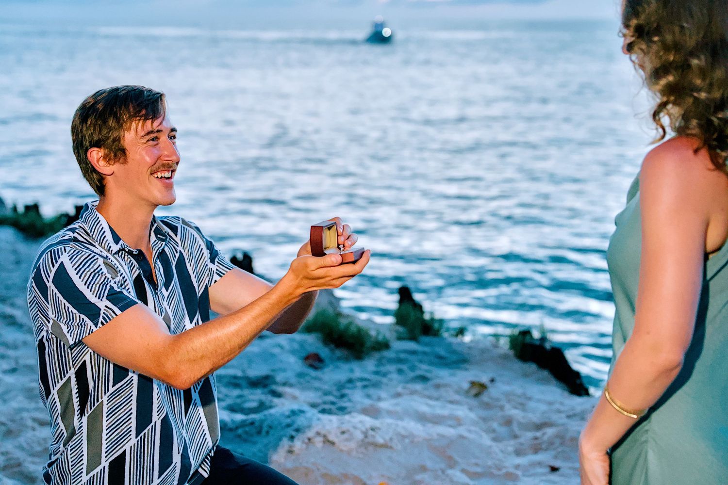 Man kneeling on beach proposing to woman with ring box at sunset by the ocean.