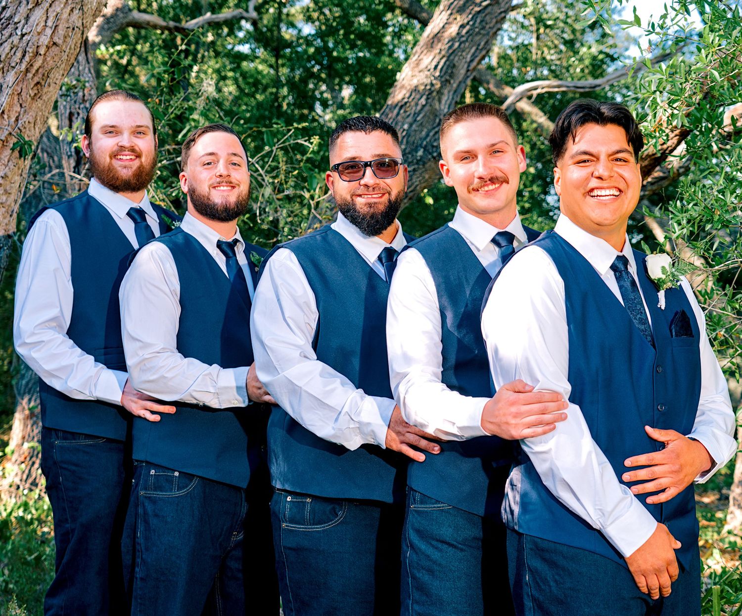 Five groomsmen in matching navy vests and white shirts posing together outdoors among trees.